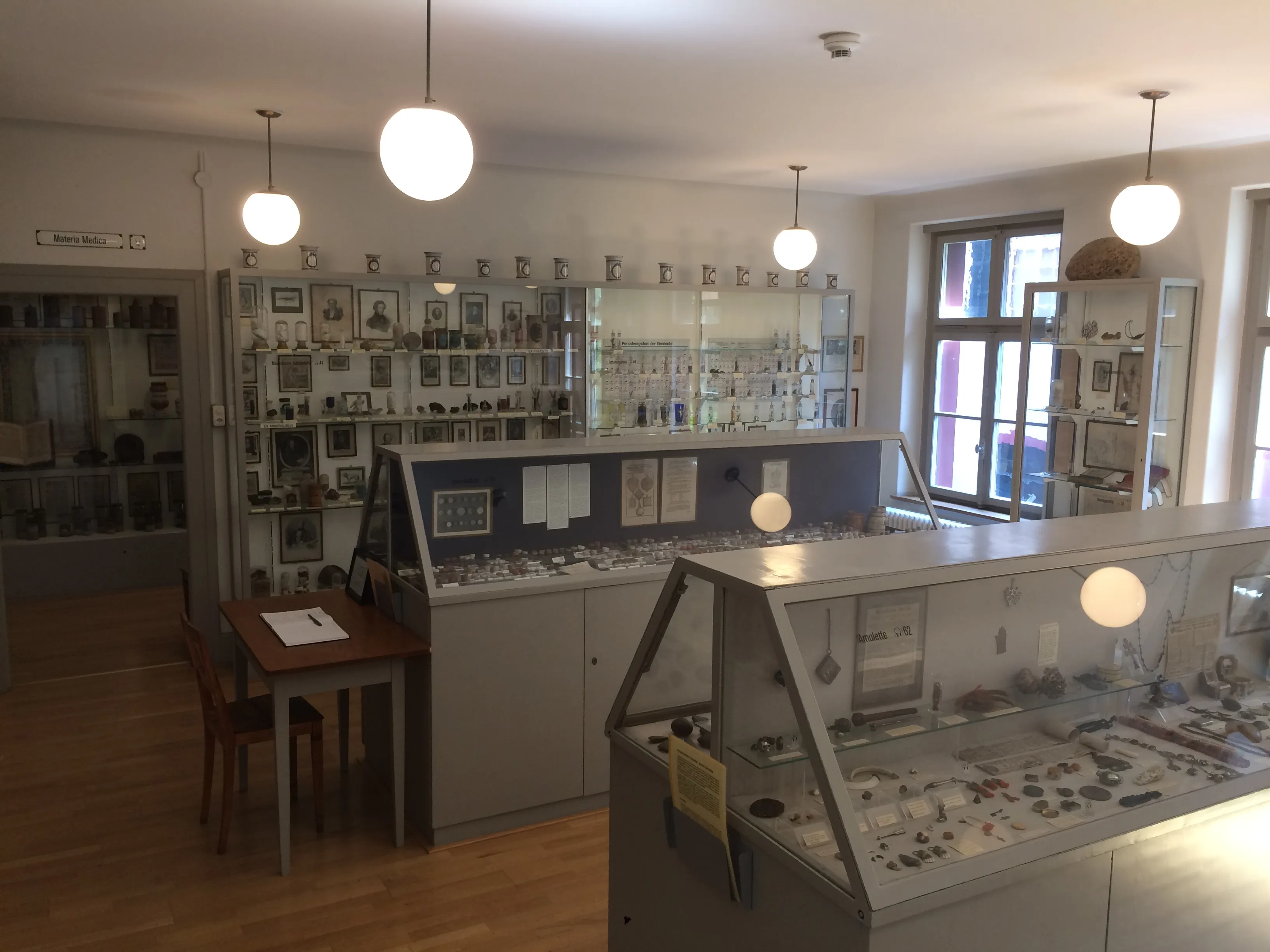 Interieur van een museumzaal met glazen vitrines met historische objecten, wandkasten en hangende bolvormige verlichting.