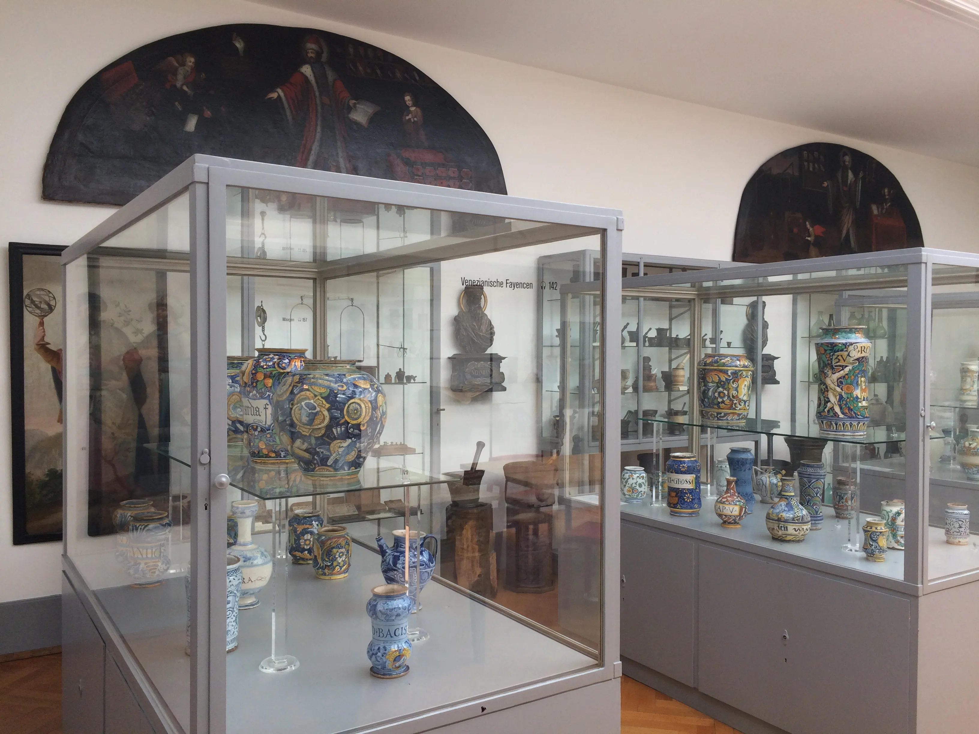 Glazen vitrines gevuld met historische keramische apothekerspotten en vaten, met op de achtergrond gewelfde schilderingen op de muur.