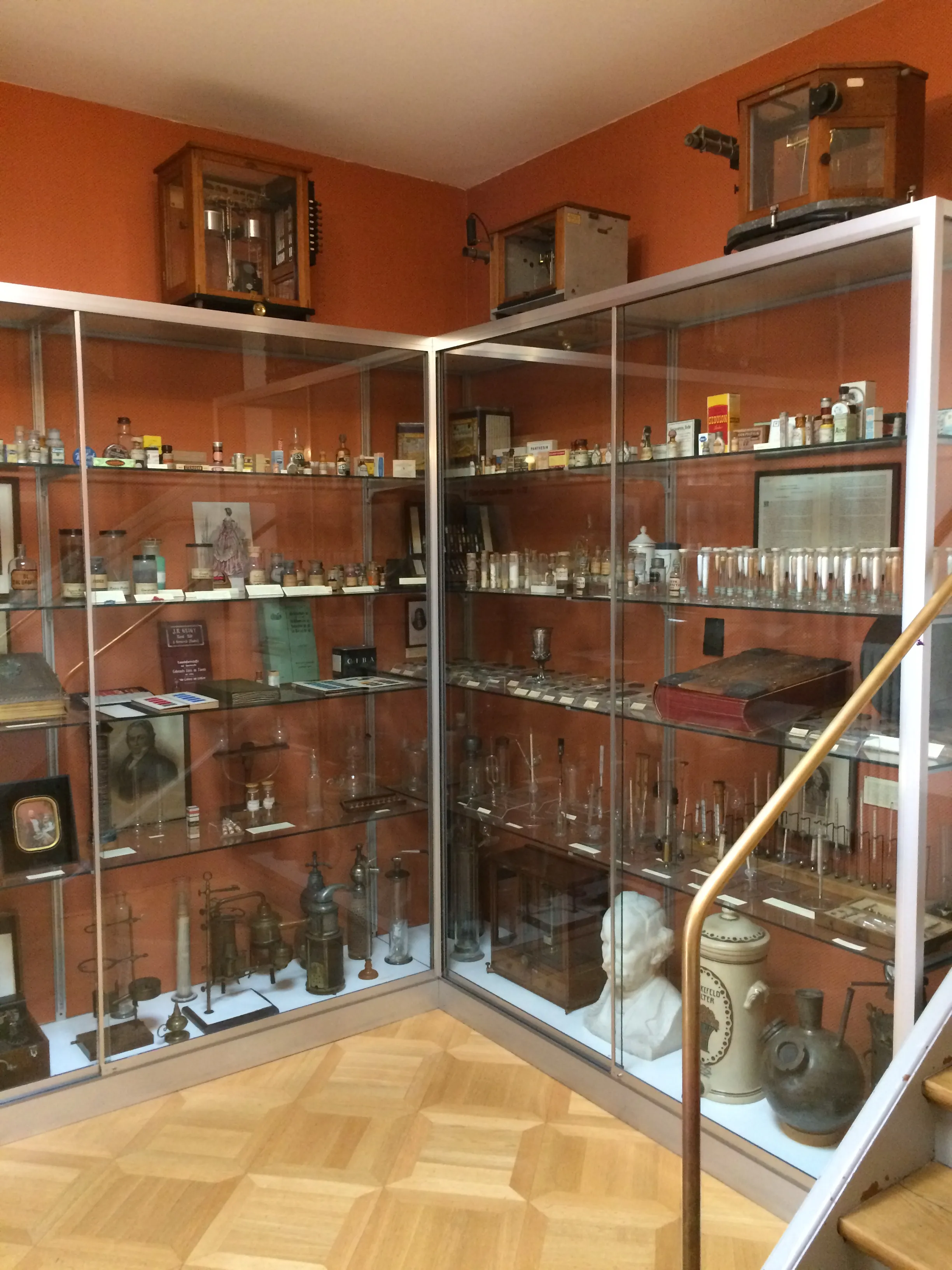 Hoek van een museumzaal met oranje muren, glazen vitrines gevuld met apothekersflessen, instrumenten en artefacten op een parketvloer, met houten kasten aan de muur erboven.