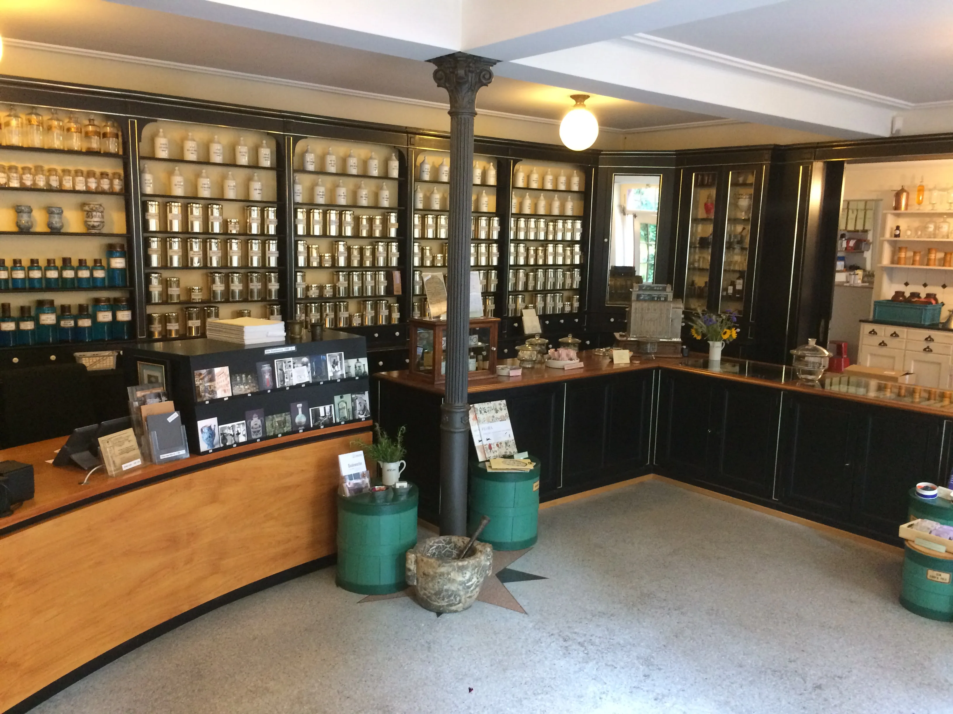 Interieur van een apothekersmuseum met wanden vol glazen apothekerspotten en flessen op donkere schappen, een gebogen houten balie, een centrale zuil en groene containers op de vloer
