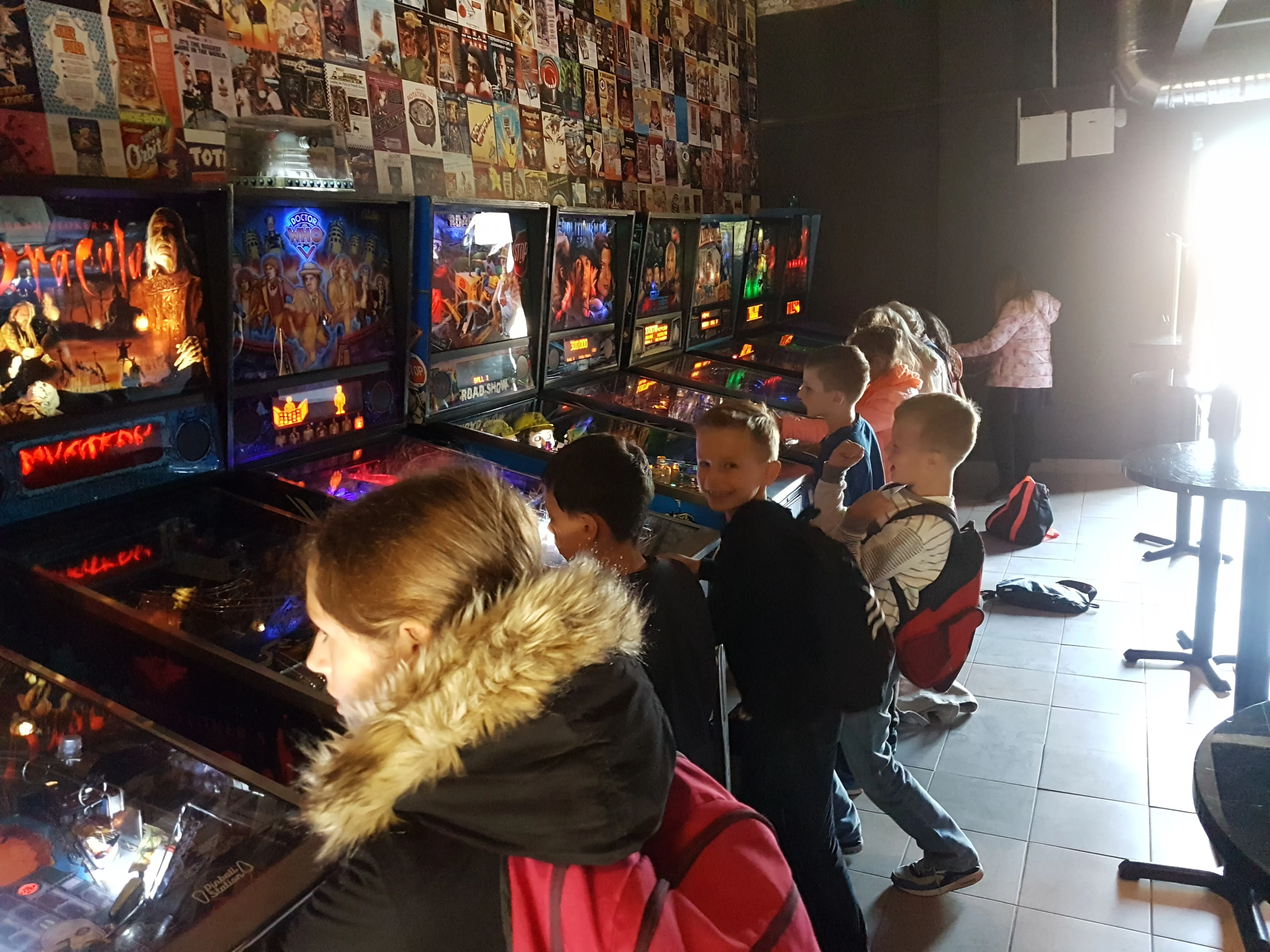 children playing pinball machines in a dimly lit arcade with colorful game screens and a wall covered in posters