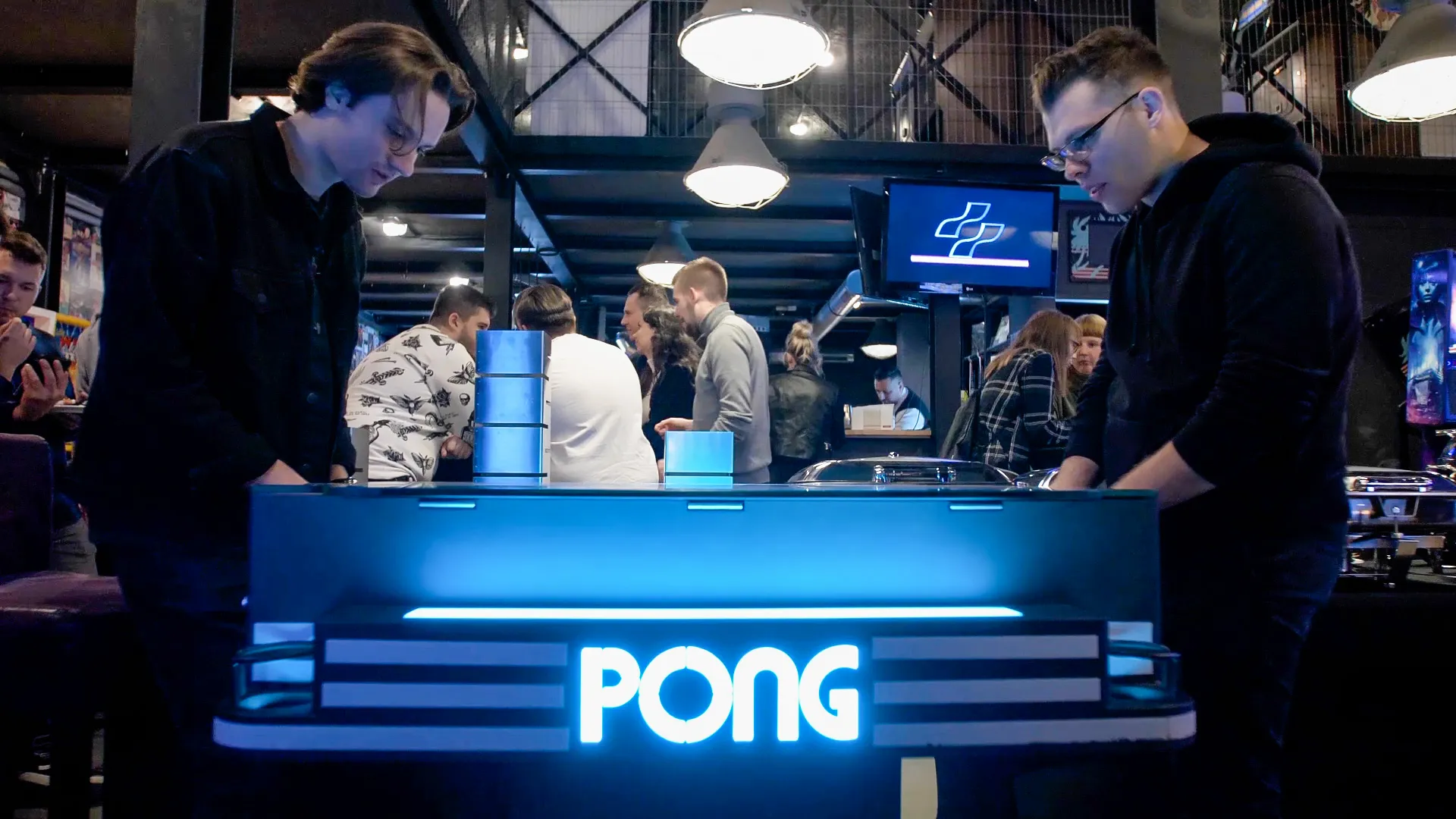 Two people playing a Pong arcade machine with a glowing blue cabinet and the word PONG illuminated in white letters