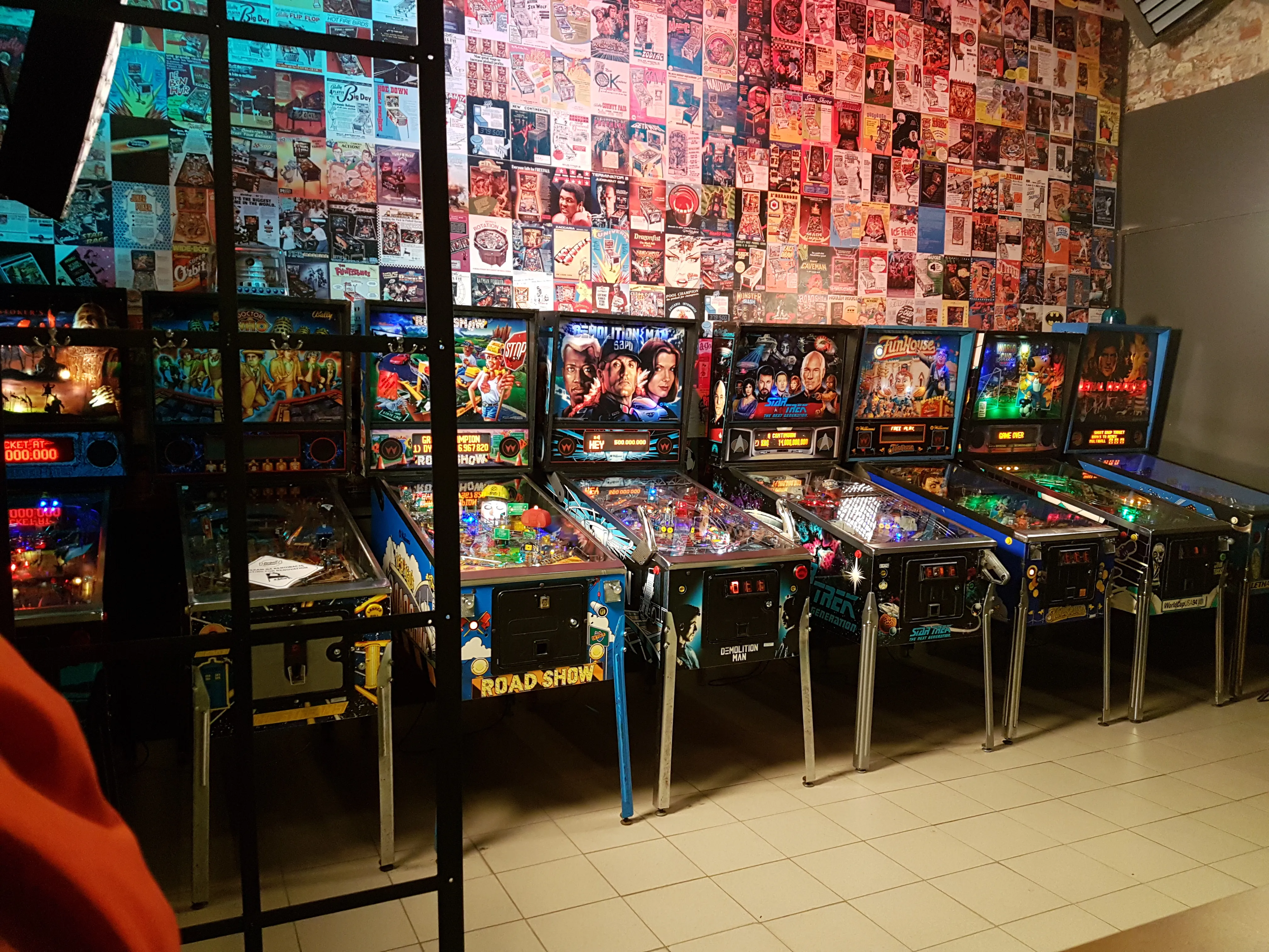 Varias máquinas de pinball alineadas en una sala de juegos con una pared de pósters de videojuegos detrás