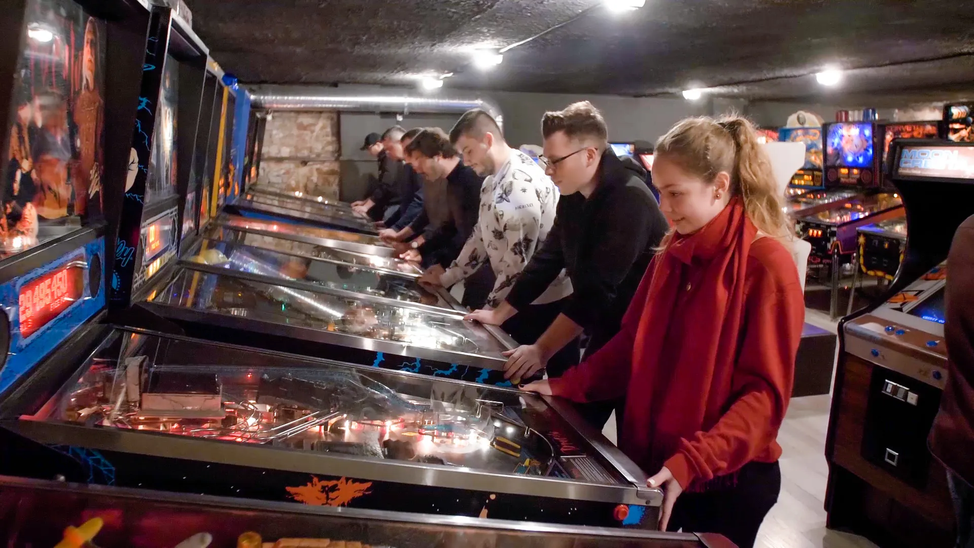 People playing vintage pinball machines in a dimly lit arcade room