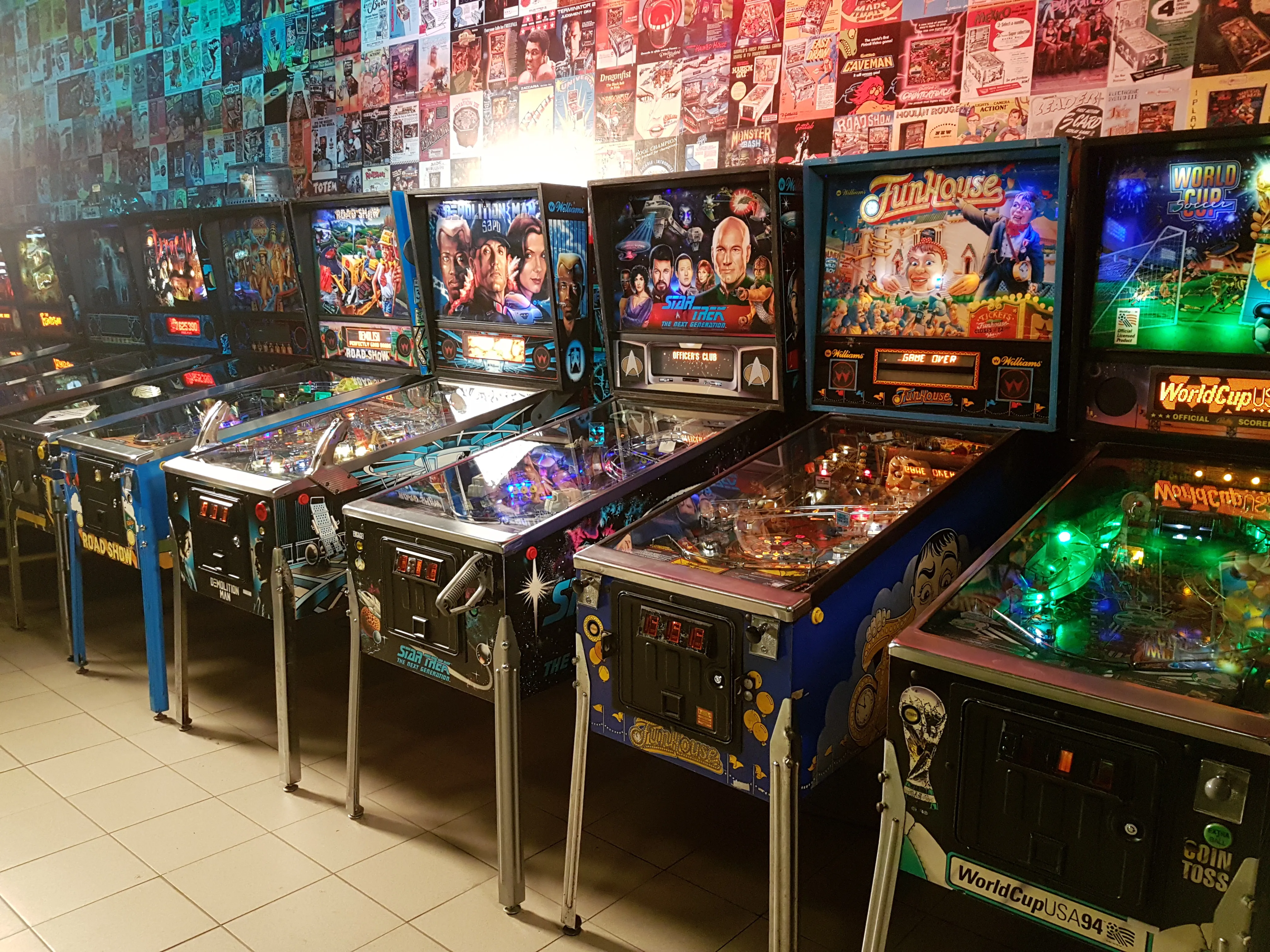 A row of vintage pinball machines with illuminated backglass displays and colorful lights in an arcade setting
