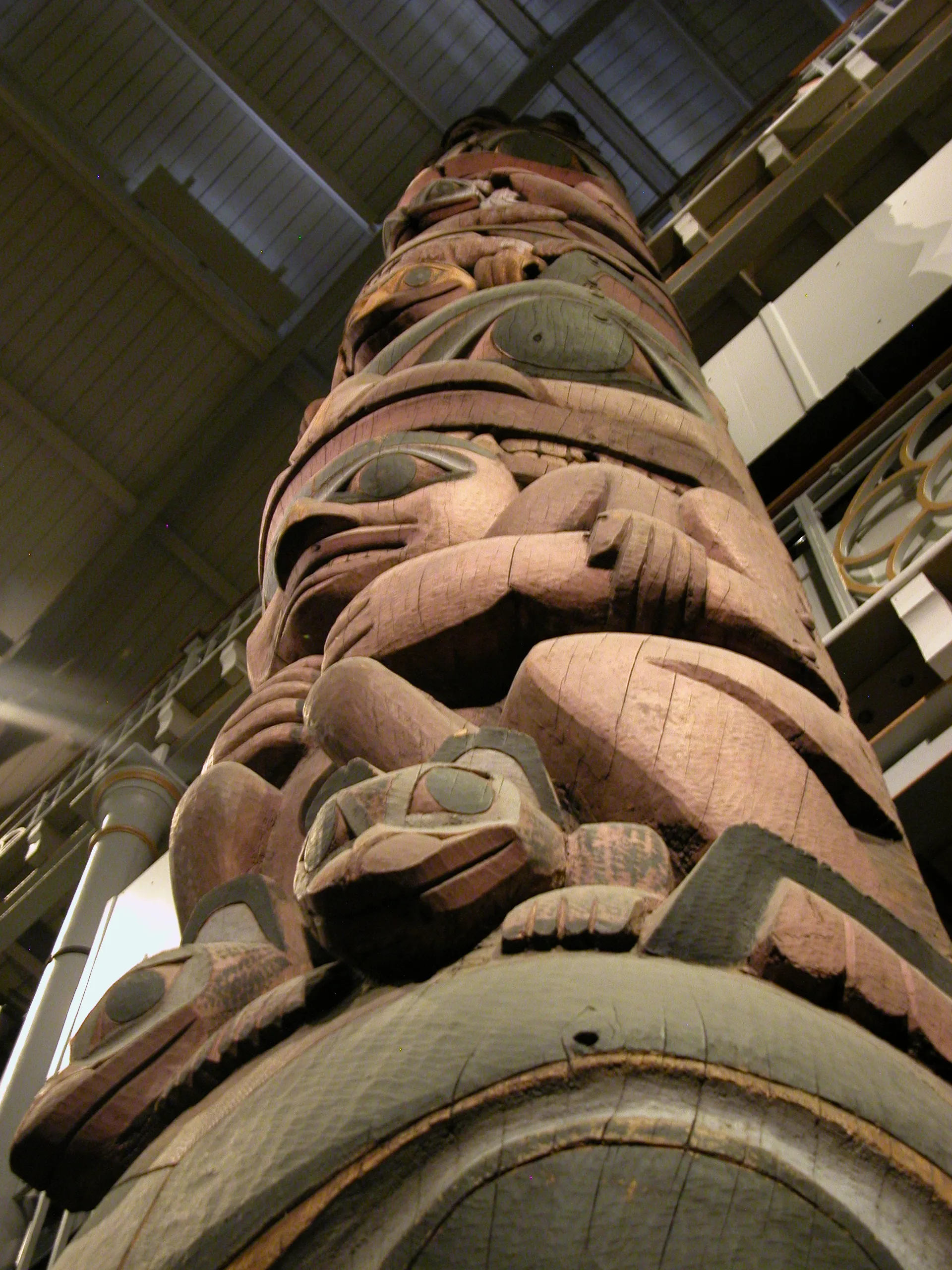 Wysoki, drewniany, rzeźbiony totem z malowanymi postaciami, widziany z dołu wewnątrz sali muzealnej, ukazujący szczegółowe, pionowo ułożone rzeźby rdzennych twórców.