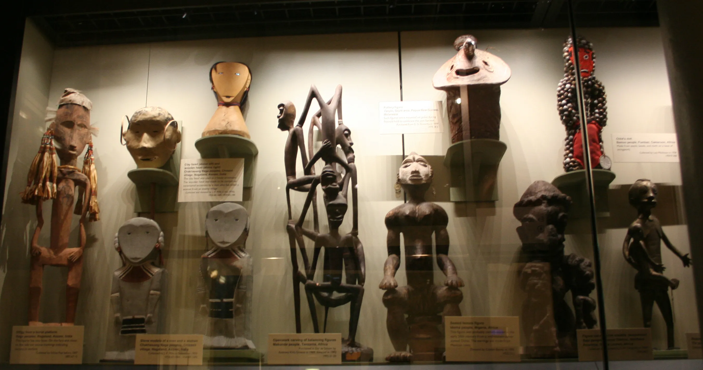 Gablota muzealna wypełniona drewnianymi rzeźbami, rzeźbionymi maskami oraz artefaktami antropologicznymi ustawionymi na półkach z tabliczkami informacyjnymi