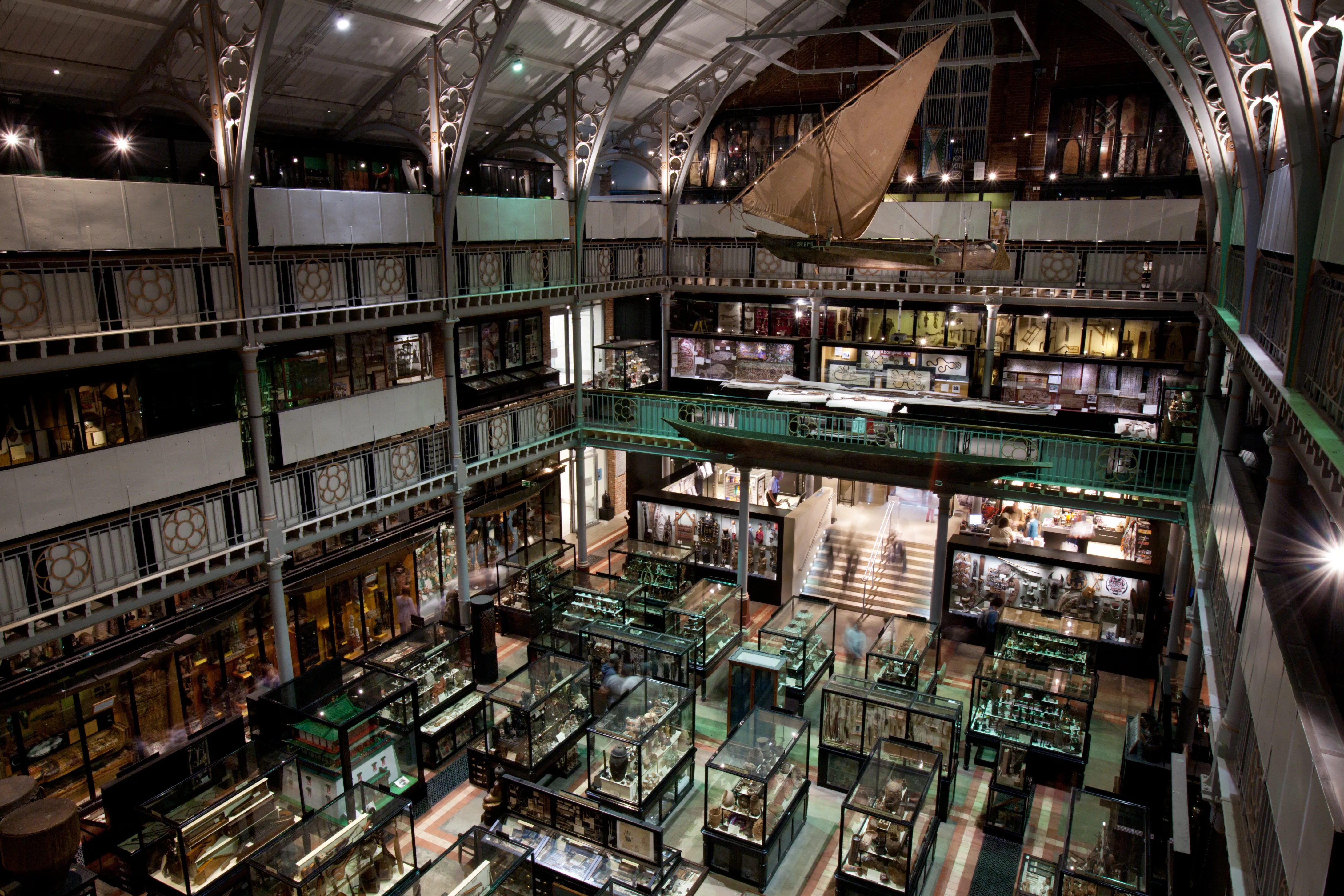 Widok z góry na wnętrze Pitt Rivers Museum, przedstawiający kilka poziomów galerii ze szklanymi gablotami, żelaznymi kolumnami i wielkim czółnem zawieszonym pod sufitem