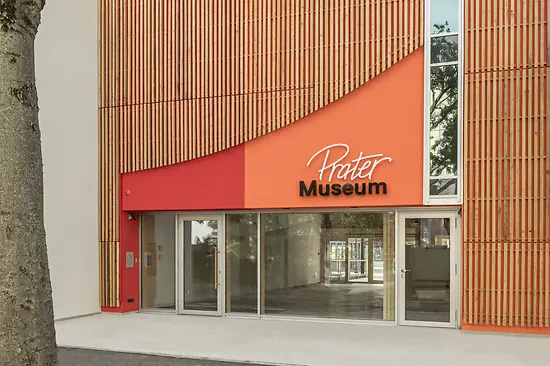 ingang van een modern gebouw met een oranje en rode gevel en het Prater Museum-bord