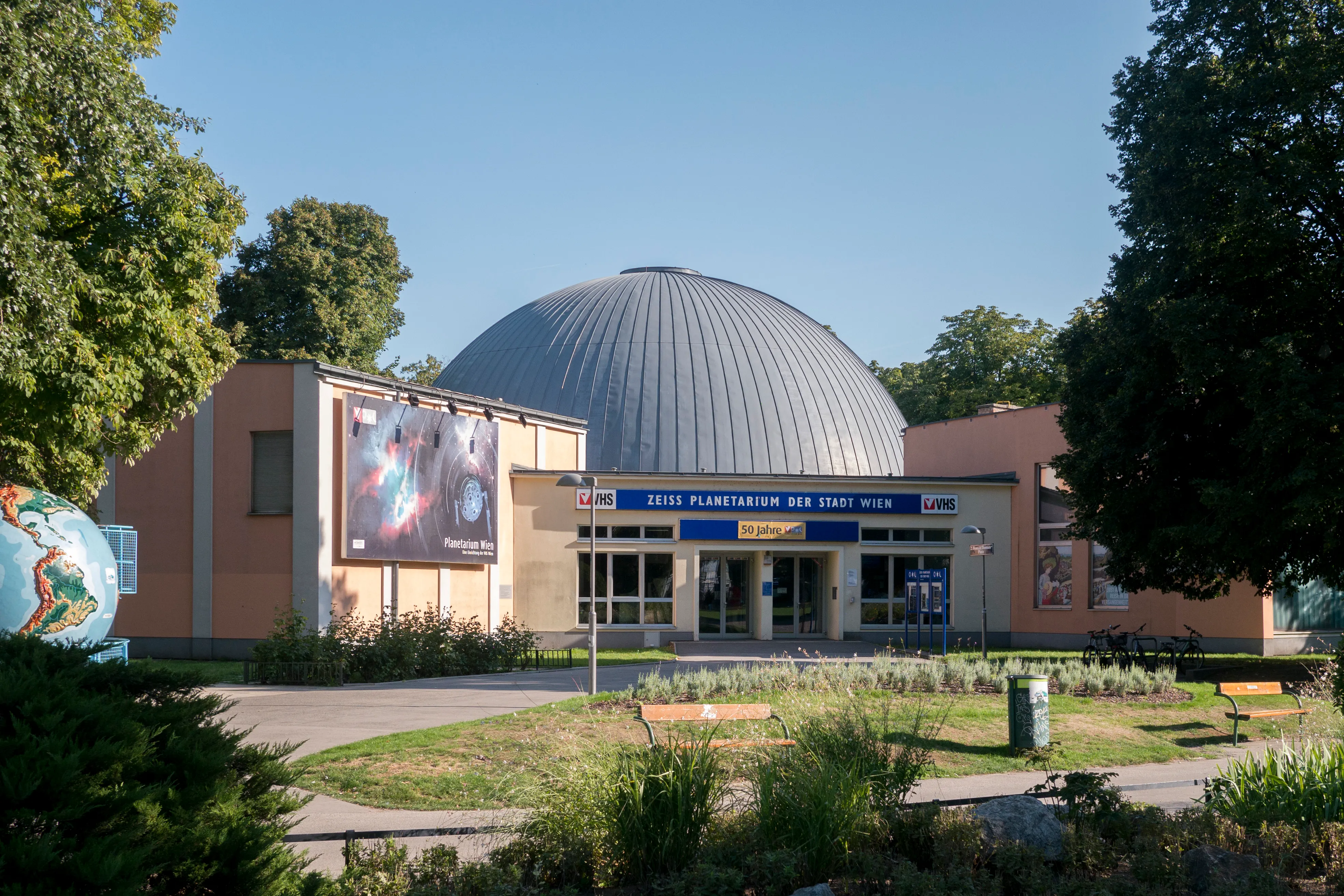 Gebouw van het Planetarium Wien met een grote koepel, ingang en omliggende groen onder een helderblauwe hemel