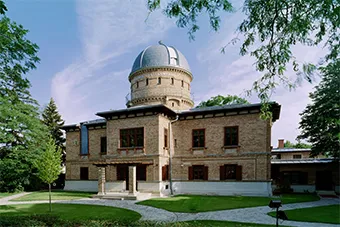 Een bakstenen gebouw met een grote koepel, omringd door bomen en een groen gazon onder een blauwe hemel.