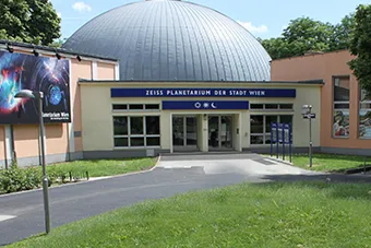Het Planetarium Wien-gebouw met een grote zilveren koepel en blauwe belettering op een beige gevel, tegen een groene grasveld en bomen.
