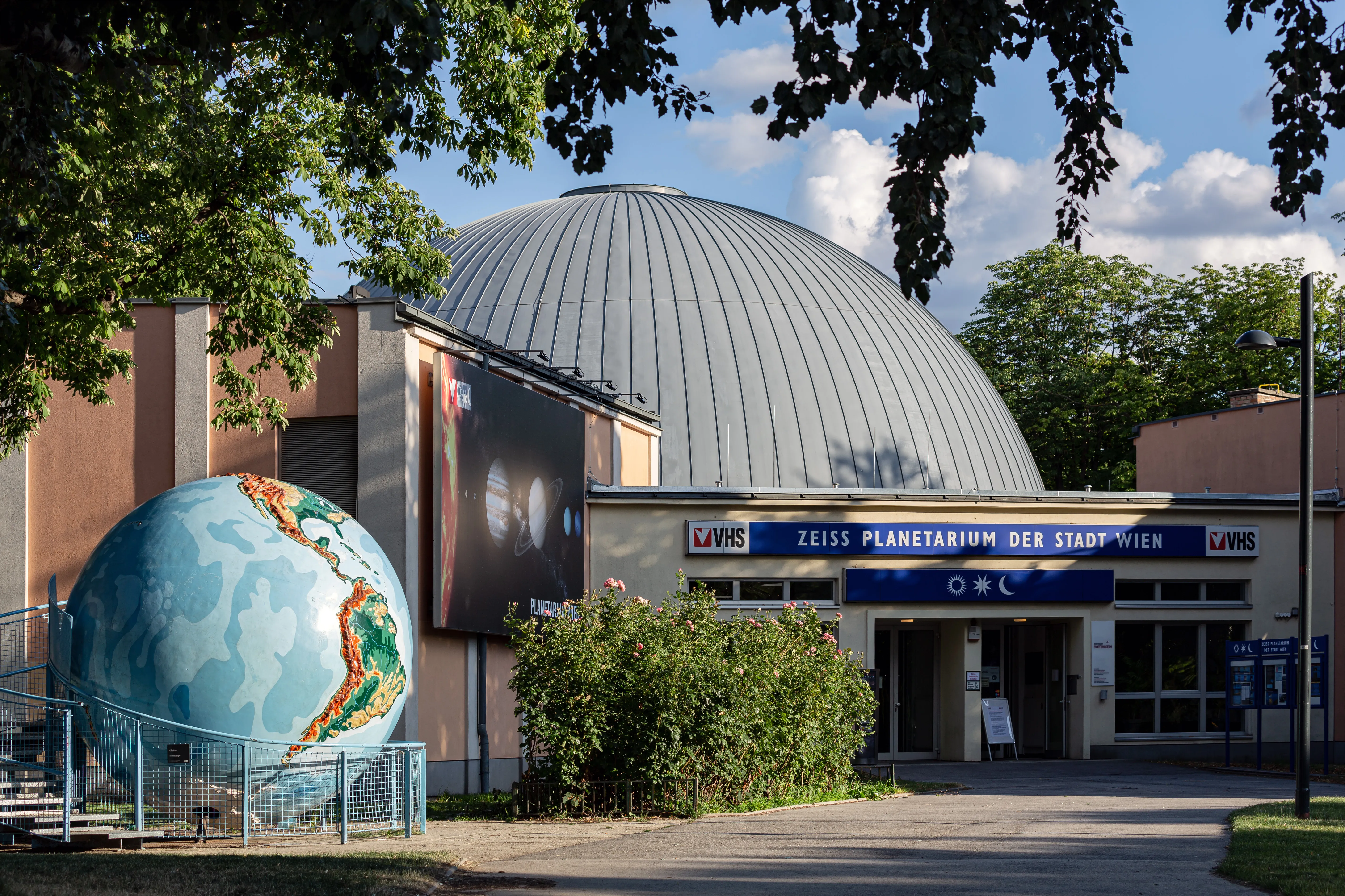 Planetarium Wien z charakterystyczną kopułą i dużą rzeźbą kuli przed nim, otoczone drzewami i zielenią.