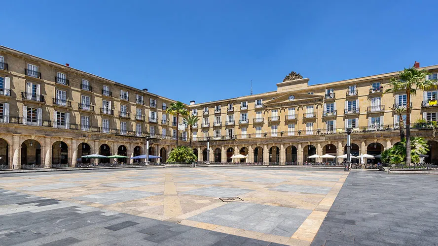 Szeroki widok na Plaza Nueva w Bilbao, przedstawiający neoklasycystyczne budynki z arkadami otaczające duży wybrukowany plac z palmami i parasolami kawiarnianymi pod bezchmurnym niebieskim niebem.