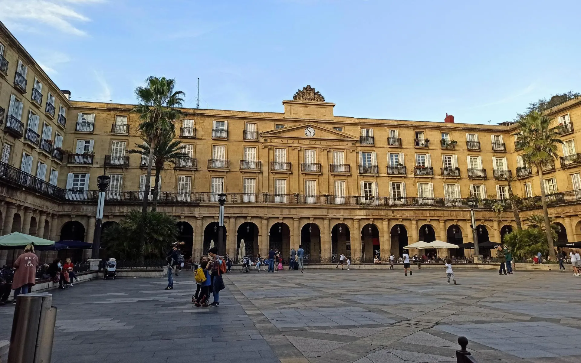 Szeroki widok na Plaza Nueva w Bilbao, ukazujący neoklasycystyczną, ochrową fasadę z arkadową kolumnadą na parterze, rzędami balkonów z żelaznymi balustradami, centralnym tympanonem z zegarem, palmami oraz ludźmi spacerującymi po brukowanym placu.