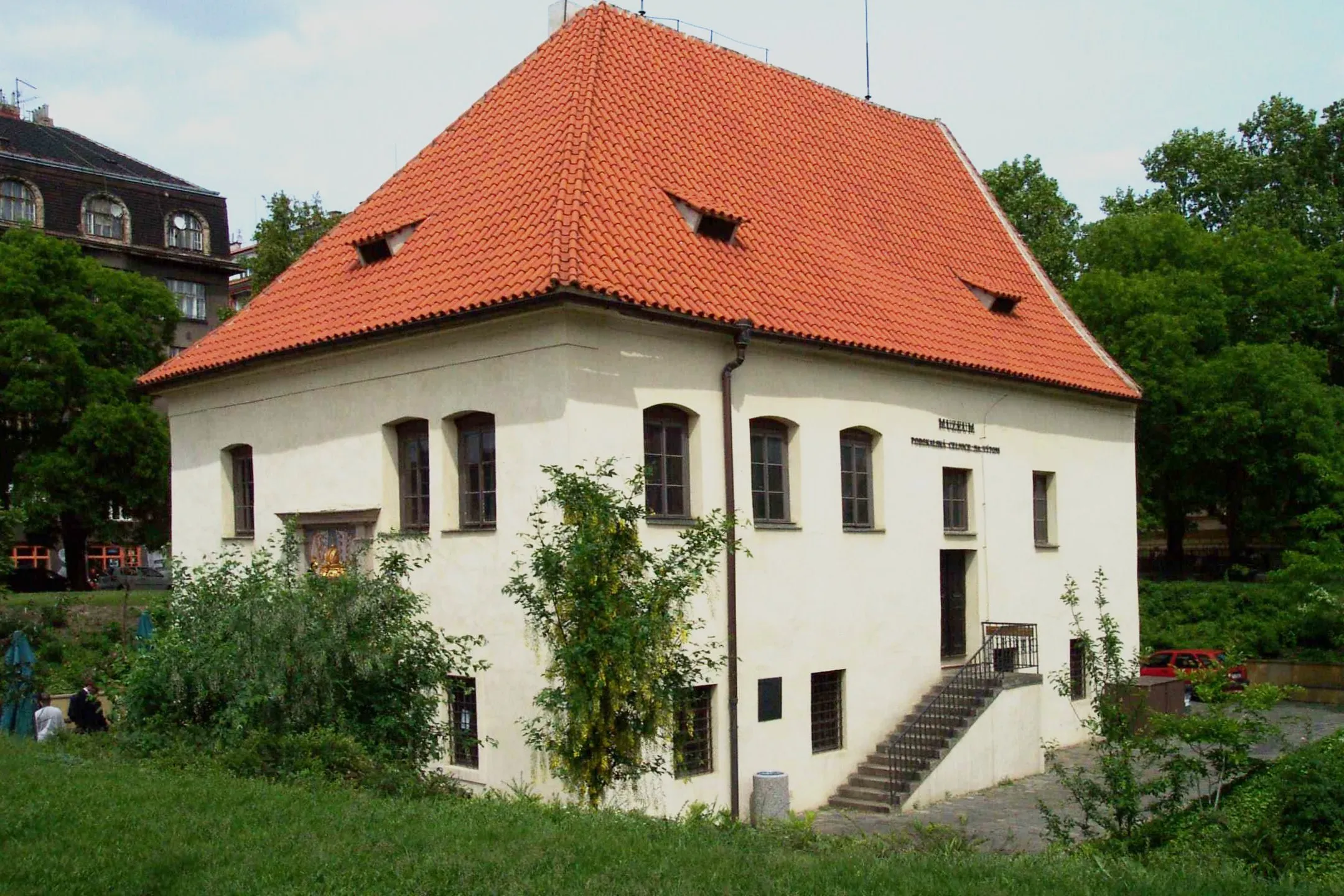 biały budynek z dachem z czerwonej dachówki i wieloma oknami, otoczony zielenią