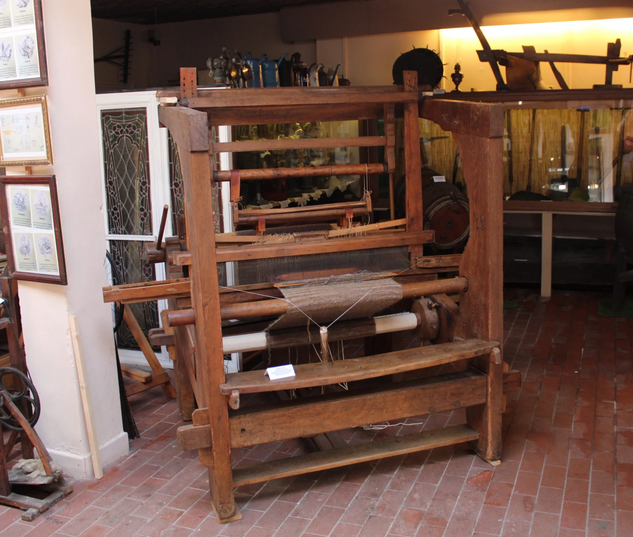 Gran telar manual de madera expuesto en una sala de museo con suelo de ladrillo y cuadros enmarcados en la pared.