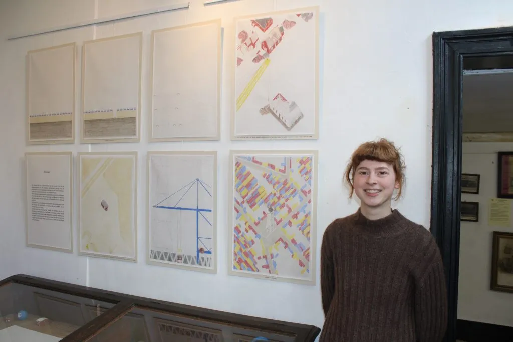Een glimlachende vrouw staat voor een witte muur met acht ingelijste kaarten en diagrammen in het Poldermuseum Lillo, met een glazen vitrinekast eronder zichtbaar.