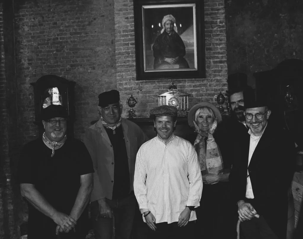 Una foto grupal en blanco y negro de seis personas sonrientes de pie en una habitación de paredes de ladrillo, con un retrato enmarcado colgado en la pared detrás de ellos.