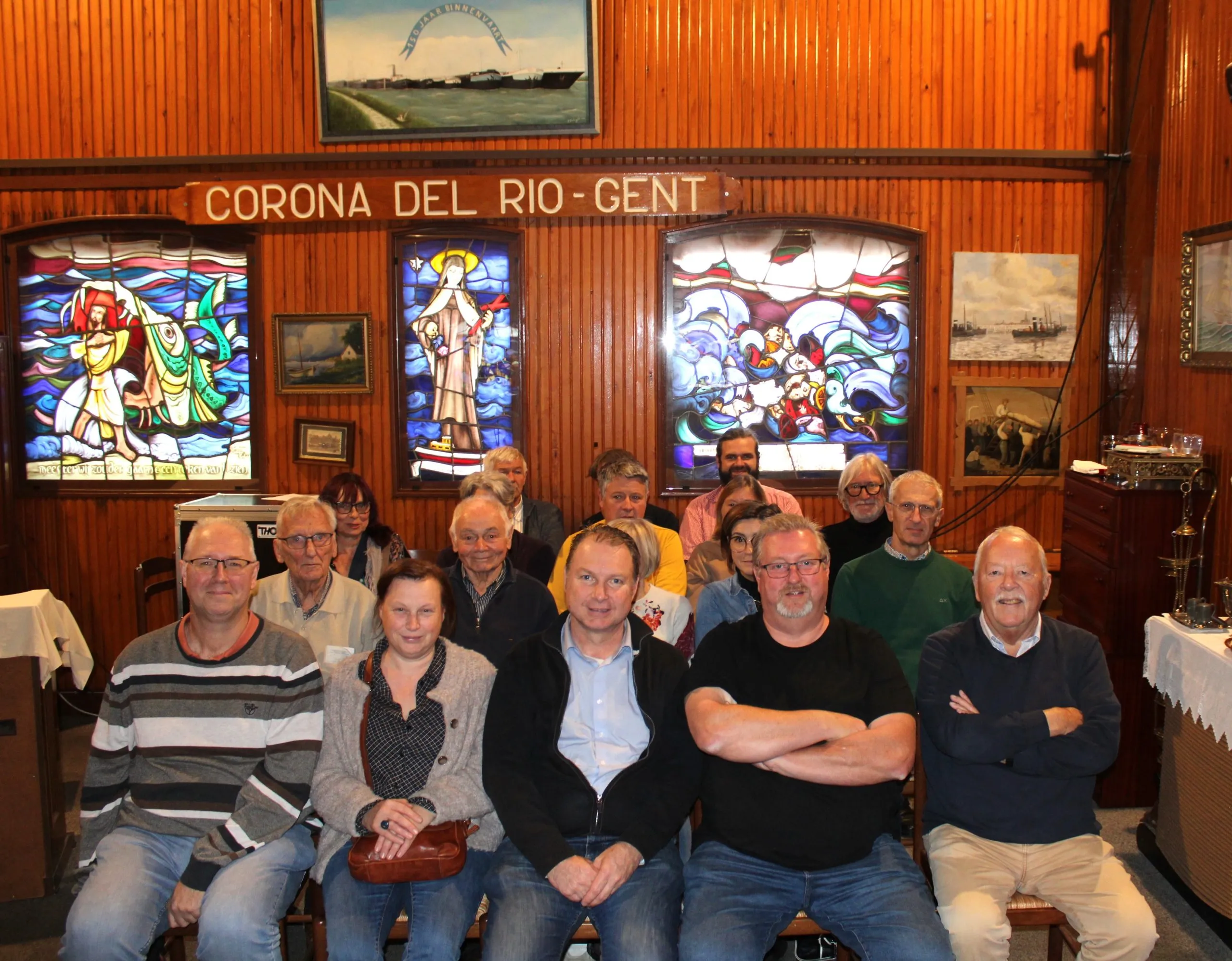 Groep mensen poserend in een kamer met houten lambrisering, glas-in-loodramen en een bord met de tekst Corona del Rio Gent