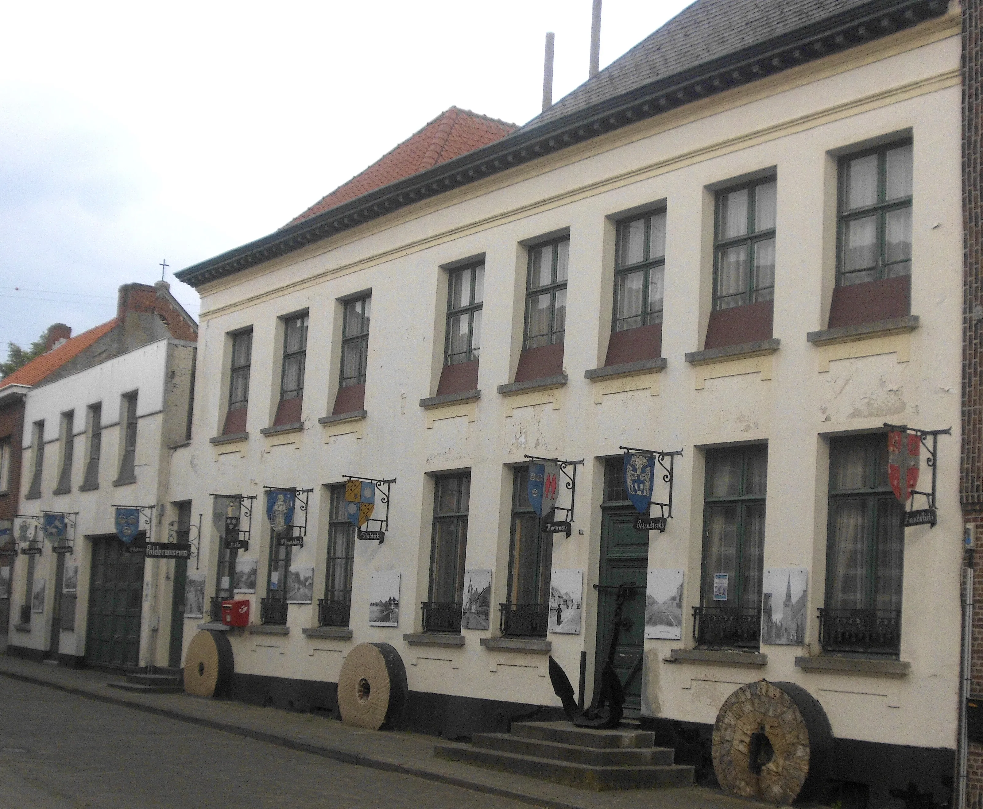 Fachada blanca de dos pisos del Poldermuseum Lillo con pancartas, piedras de molino y un ancla expuestos en el exterior