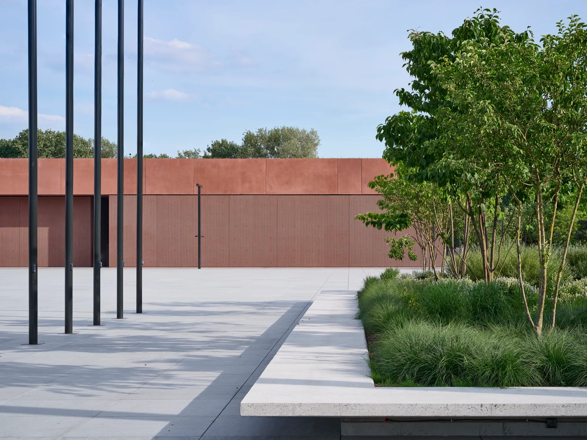 modern gebouw met roestkleurige gevel, betonnen looppad en verticale zwarte palen omringd door groene planten