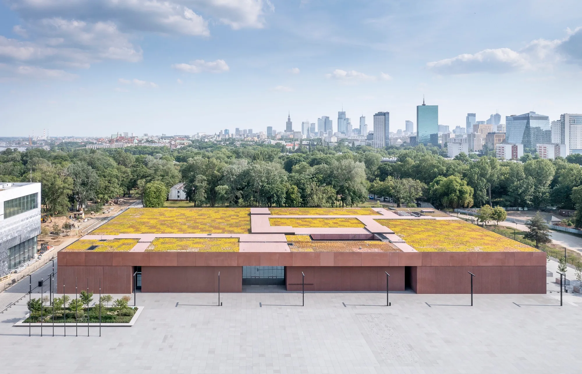 Modern gebouw met een groen dak en bruine gevel, omringd door bomen en een stadsgezicht op de achtergrond