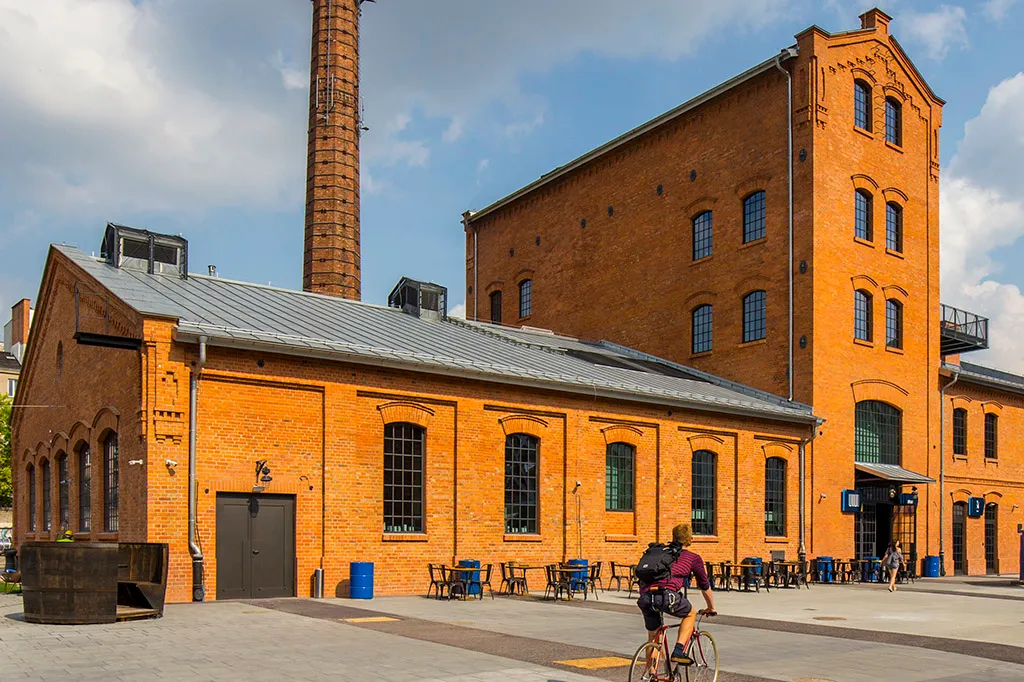 Edificio industrial de ladrillo rojo con una chimenea alta y grandes ventanales, con asientos al aire libre y un ciclista delante