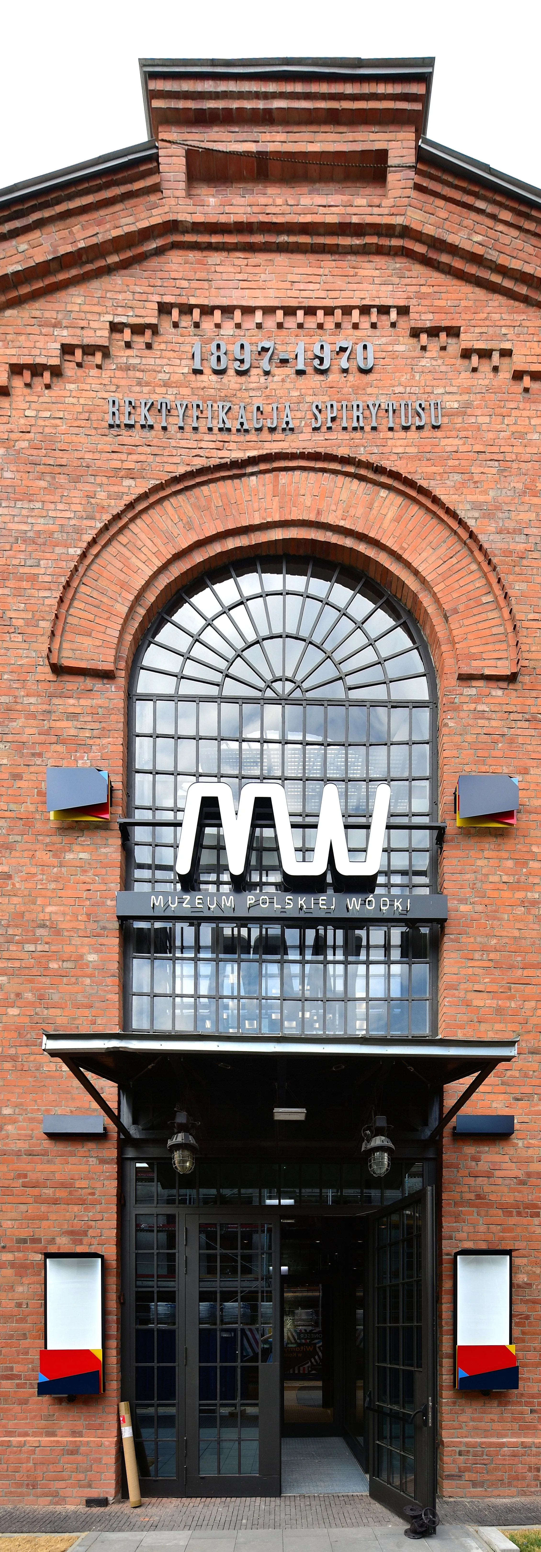 Entrada de edificio de ladrillo con ventana arqueada, que presenta el letrero 'MUZEUM POLSKIEJ WÓDKI' y un gran logotipo 'MW' encima de las puertas.