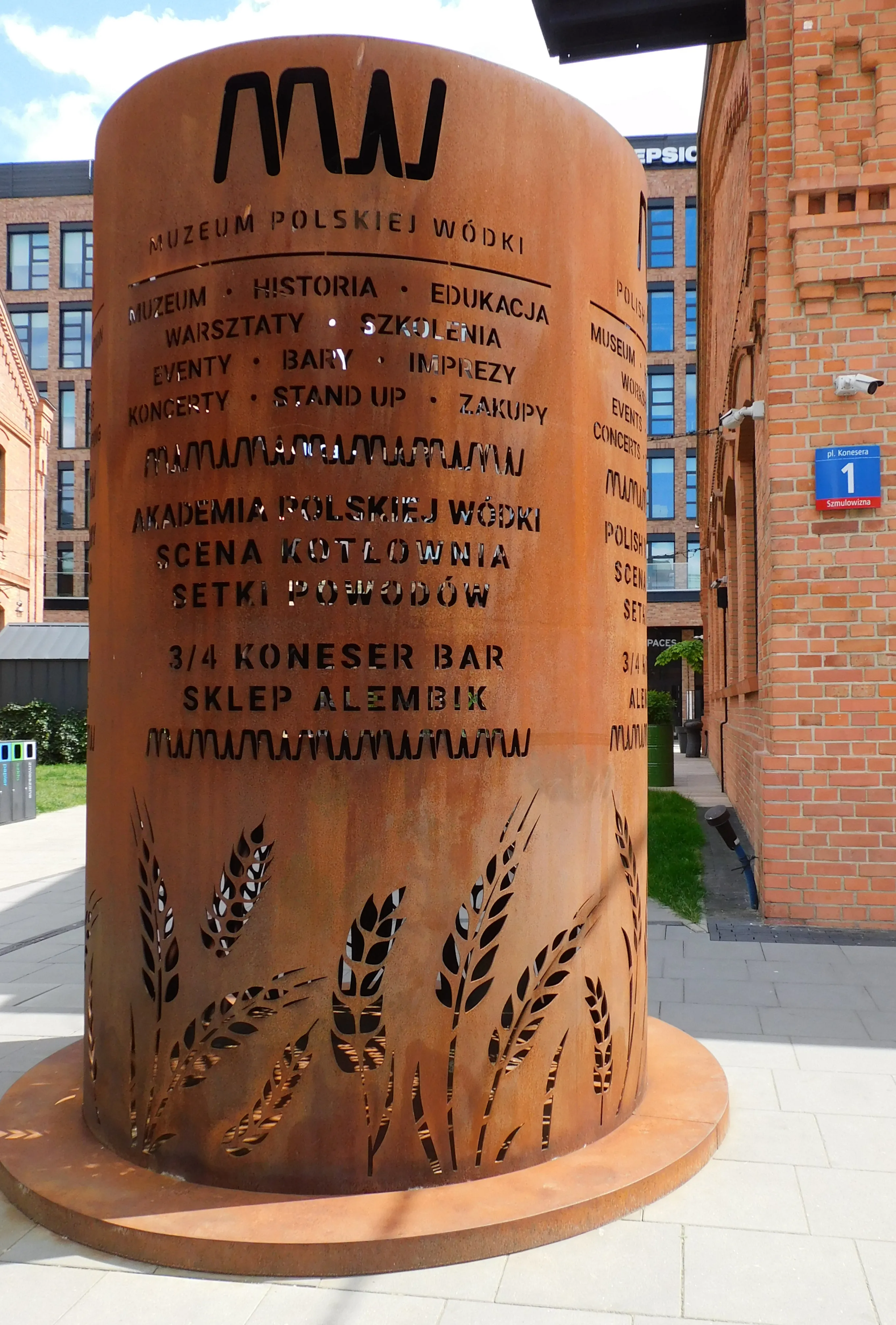 Cartel cilíndrico de color óxido para el Museo de Vodka Polaco con texto y diseños de trigo, ubicado al aire libre cerca de un edificio de ladrillos