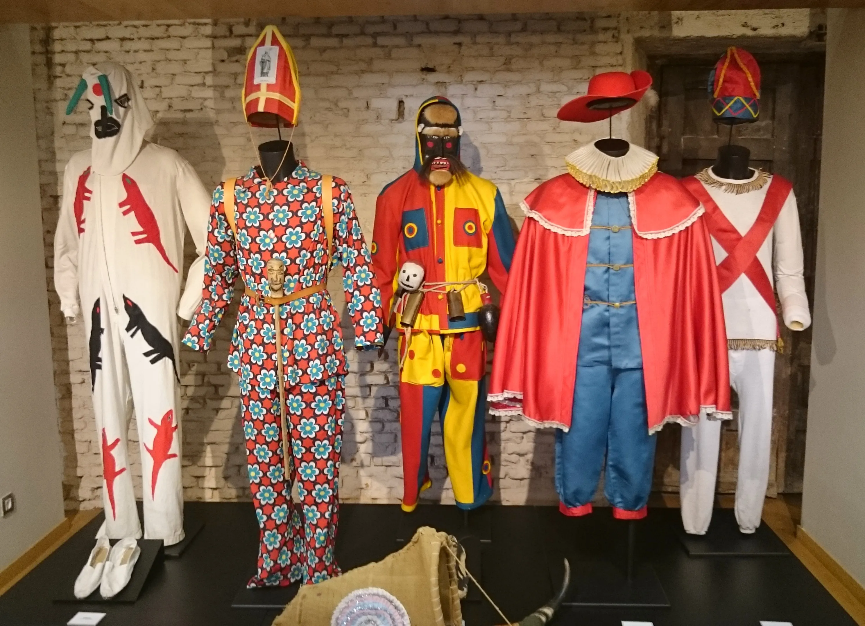 Pięć tradycyjnych strojów na manekinach, prezentujących żywe wzory i ceremonialne nakrycia głowy, wyeksponowane na tle ceglanej ściany.