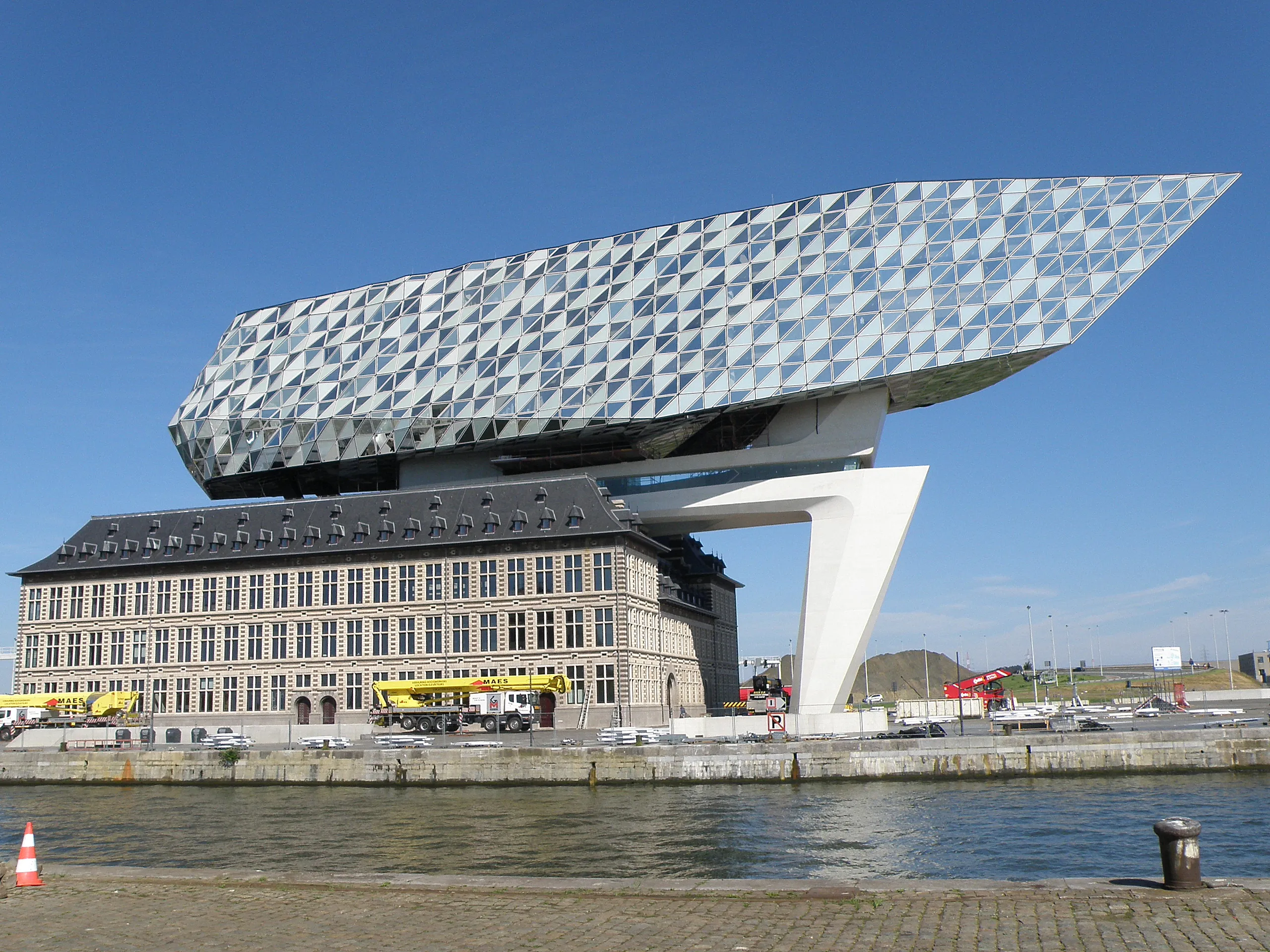 Extensión de cristal cristalino de la Casa del Puerto de Amberes que se eleva sobre un histórico edificio de ladrillo a lo largo del paseo marítimo bajo un cielo azul despejado