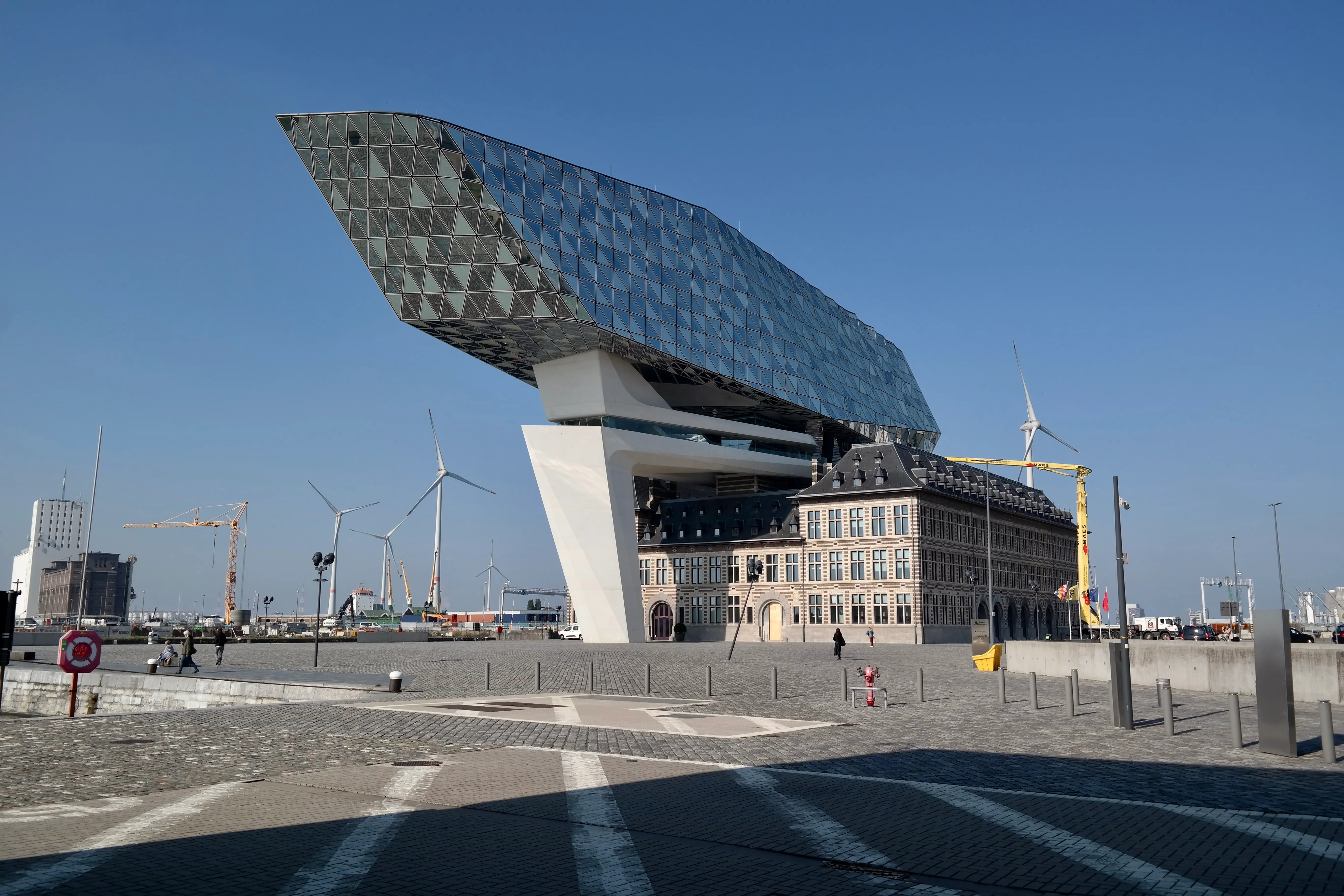 Vista exterior de la Casa del Puerto de Amberes que muestra la estructura superior angular revestida de cristal en voladizo sobre el histórico edificio de ladrillo, con aerogeneradores y una plaza pavimentada en primer plano bajo un cielo azul despejado.