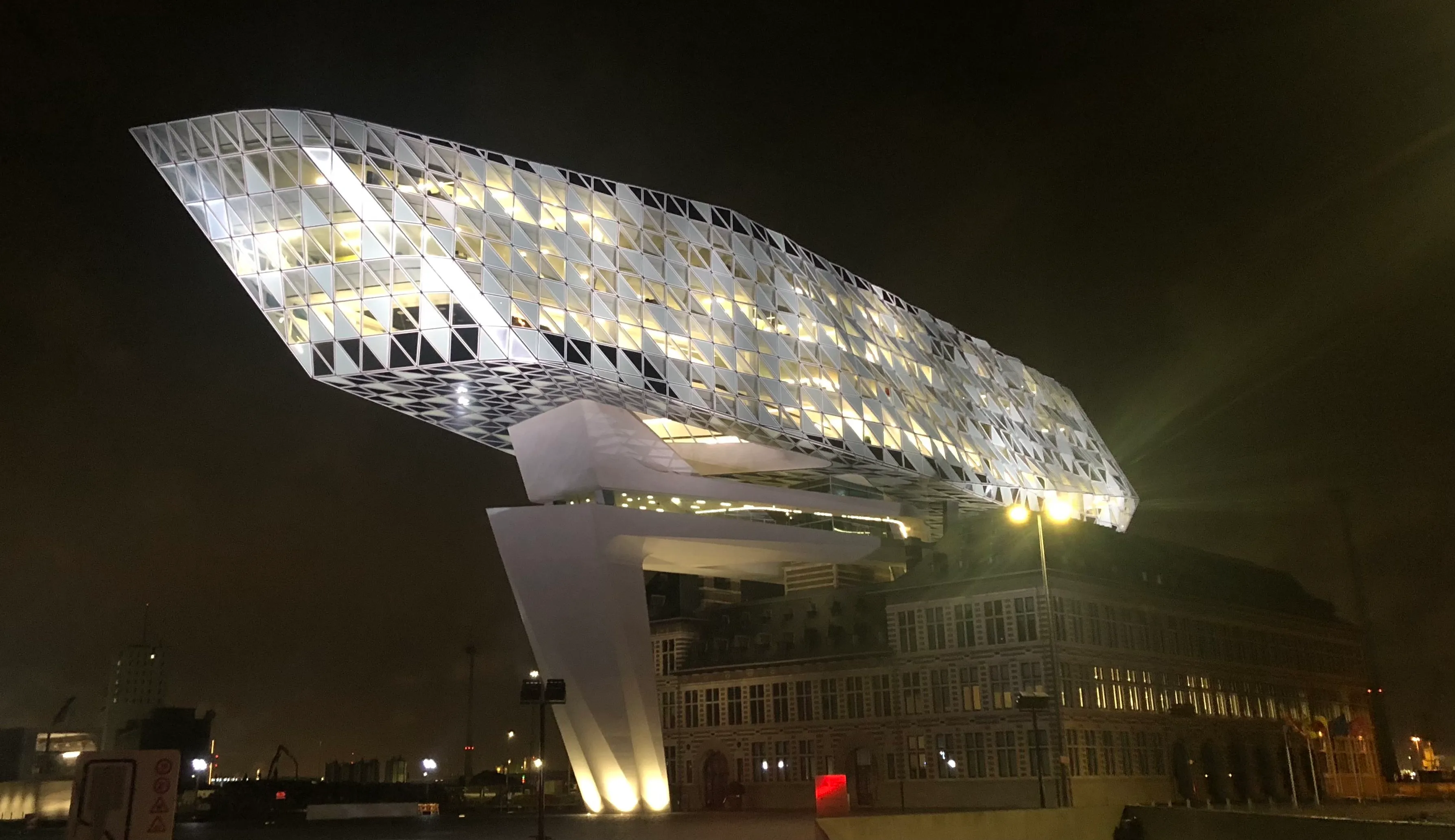 La extensión de vidrio cristalino iluminada de la Casa del Puerto de Amberes brillando por la noche sobre el histórico edificio de la estación de bomberos de ladrillo