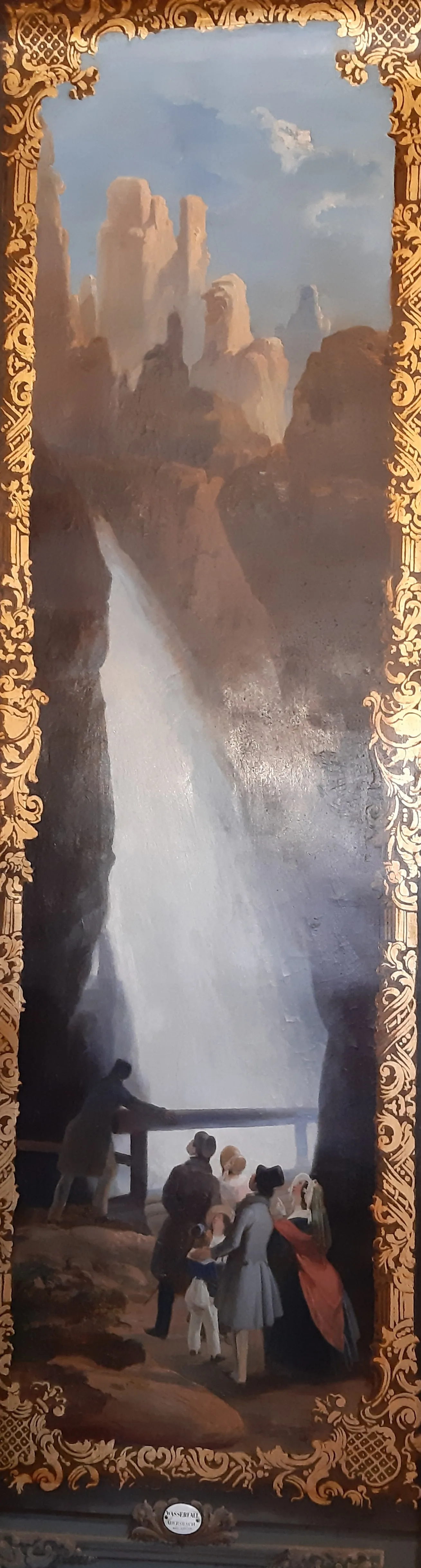 A framed painting of people viewing a waterfall in a mountainous landscape