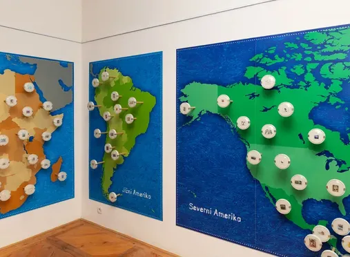 Three large world maps on a wall, each with circular objects attached, in a museum gallery.