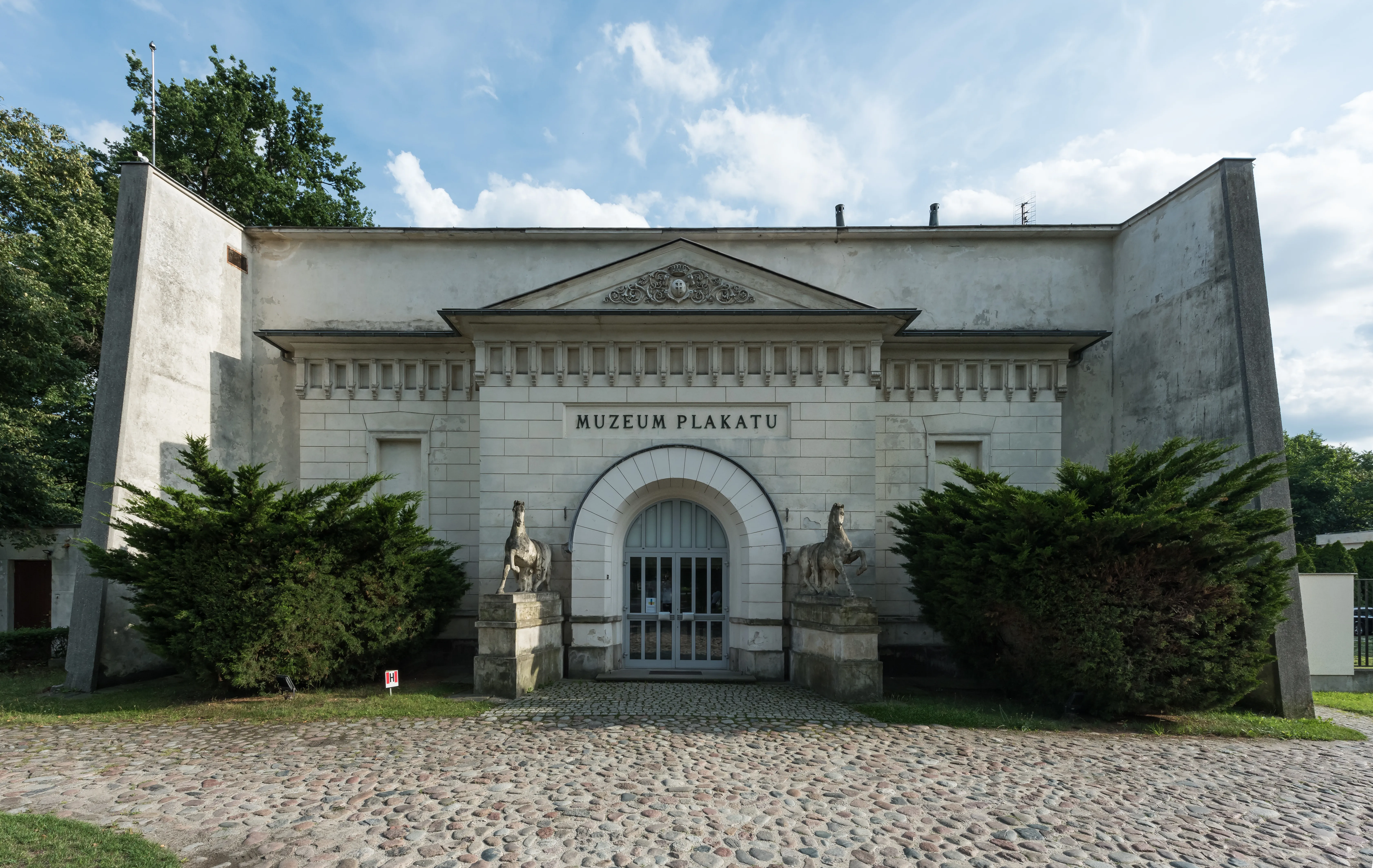 The exterior of the Poster Museum at Wilanów, showing a stone building with an arched entrance, two statues, and the name 'MUZEUM PLAKATU' above the door.