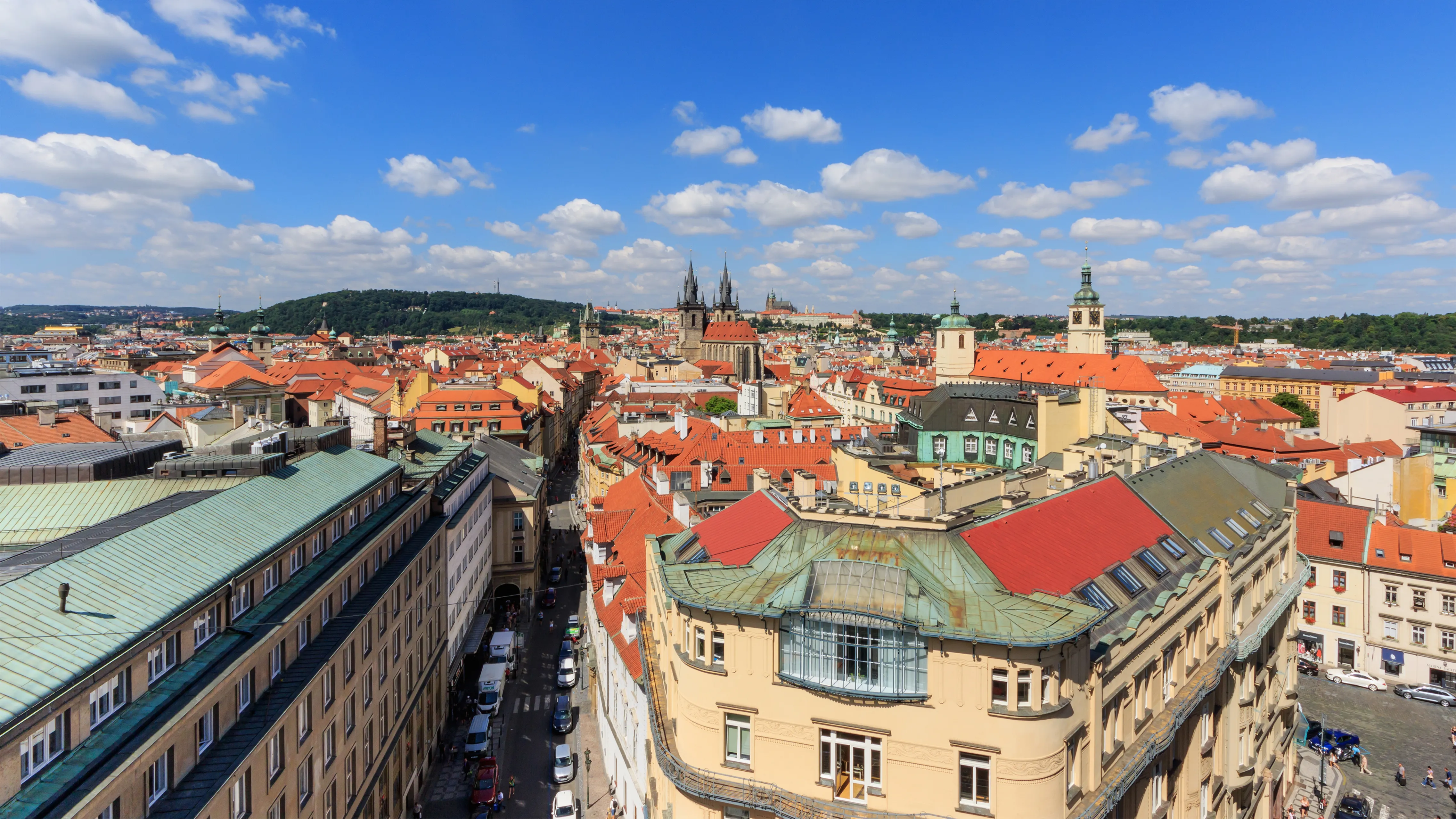 Panoramisch stadsgezicht van Praag met rood-gedekte gebouwen, een prominente toren en een blauwe lucht met verspreide wolken