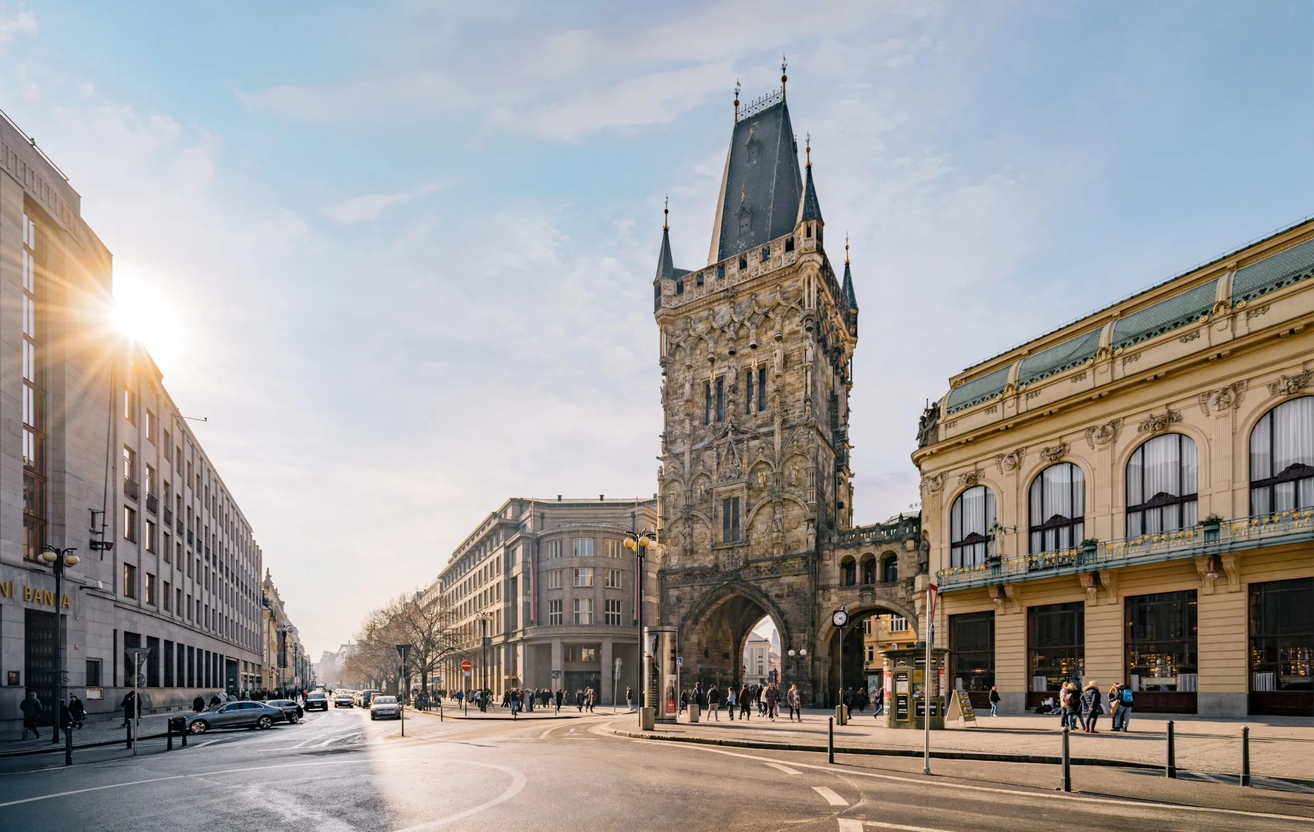 Wieża Prochowa, zabytkowa gotycka kamienna brama z wysoką iglicą, stoi na miejskim skrzyżowaniu z budynkami po obu stronach i czystym niebem nad nią.