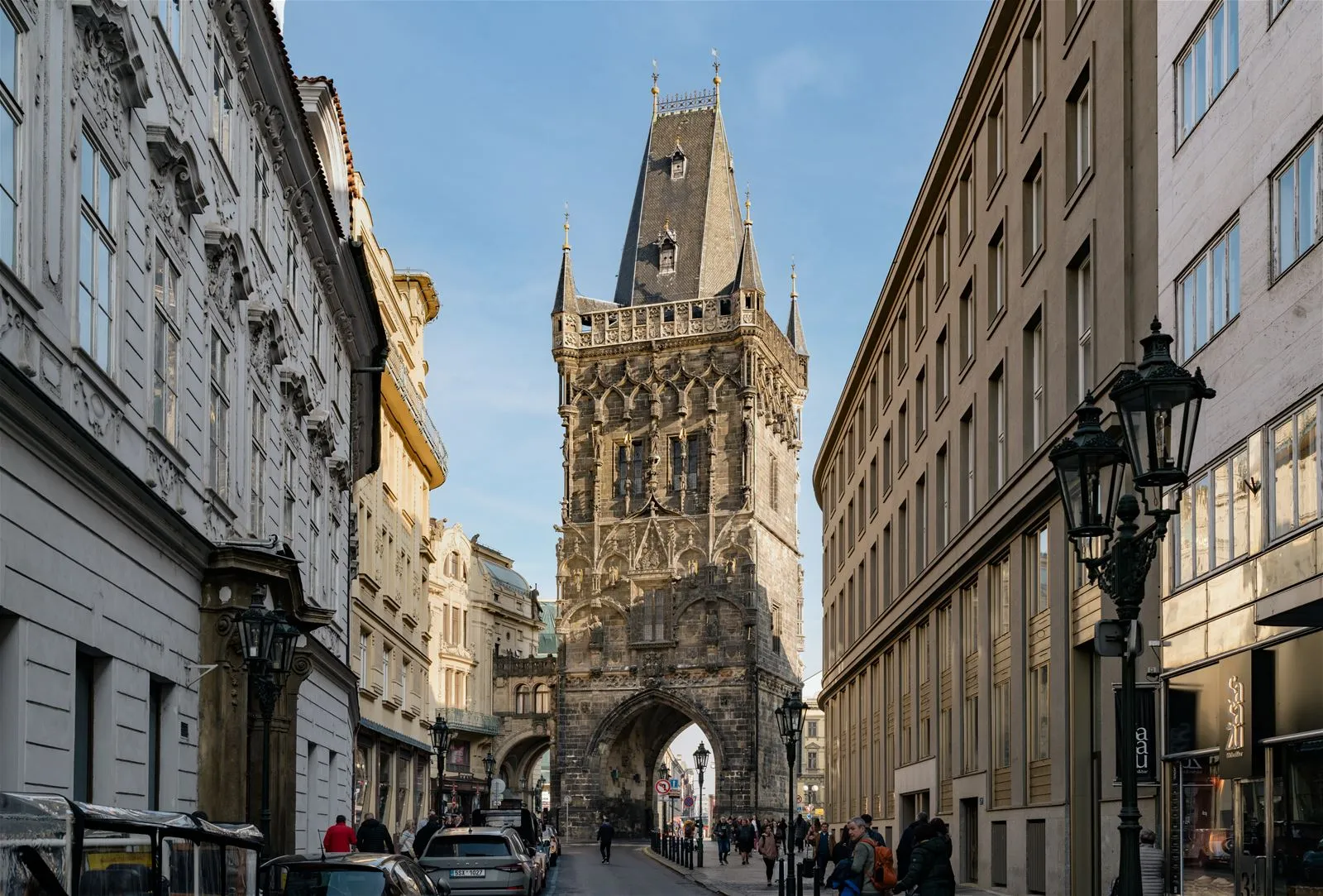 Een hoge stenen toren met een puntig dak en een boogvormige toegangspoort, gelegen tussen gebouwen aan een straat in Praag.