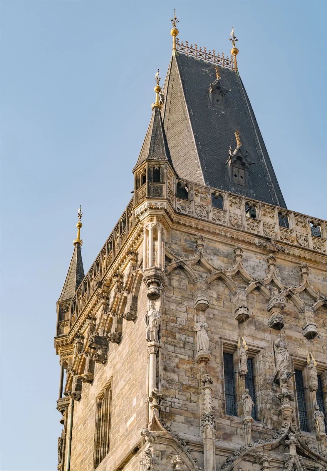 Stenen toren met sierlijke gotische architectuur, spitsen en een donker dak tegen een blauwe hemel