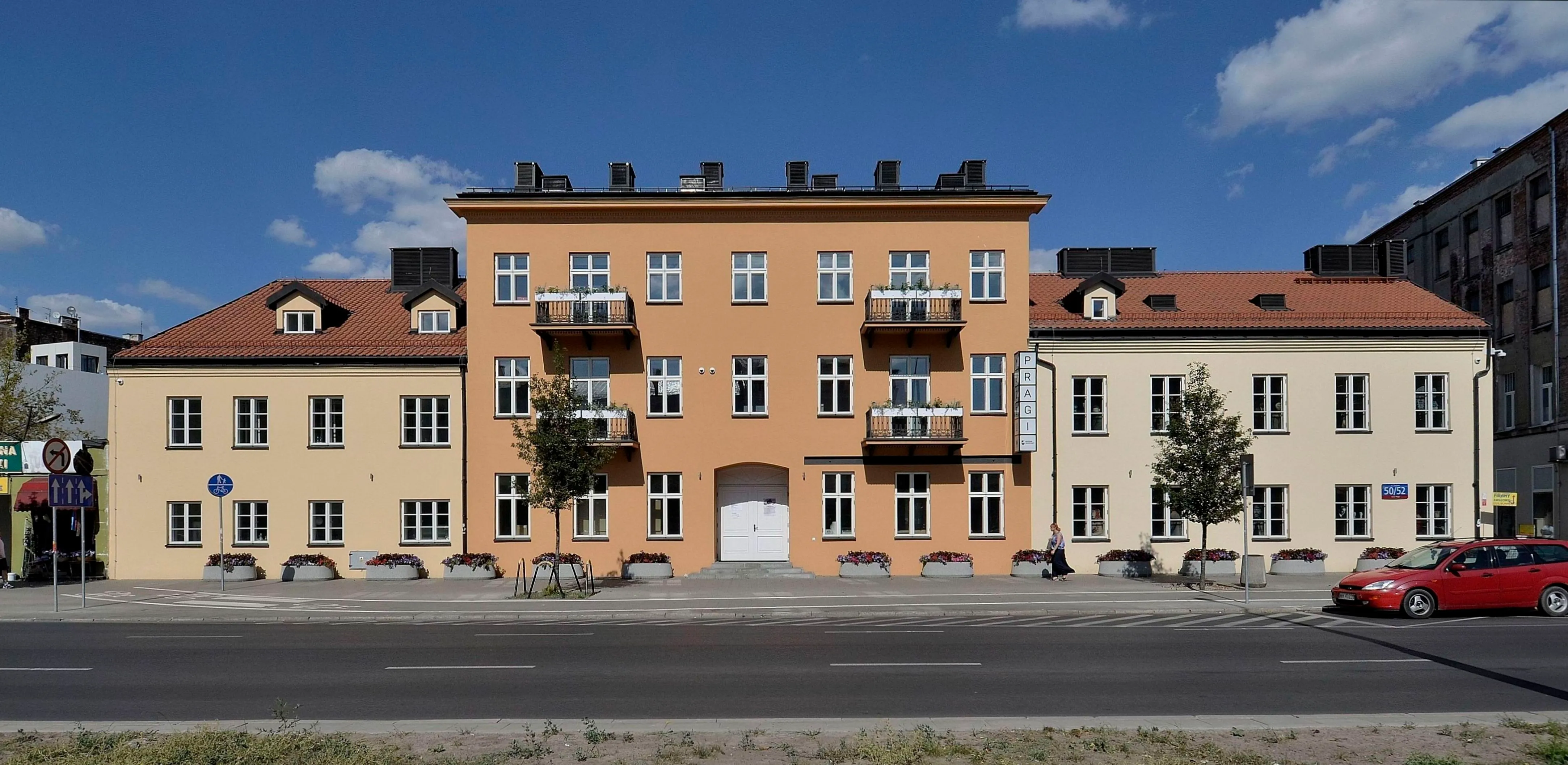 Pomarańczowo-beżowy budynek z białymi oknami i balkonami, czerwony samochód zaparkowany w pobliżu, pod błękitnym niebem
