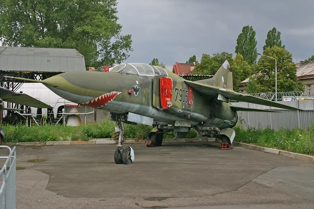 Een groene militaire straaljager met haaienbek-neuskunst en rode markeringen aan de zijkant, geparkeerd buiten op een verhard gebied met bomen en gebouwen op de achtergrond.
