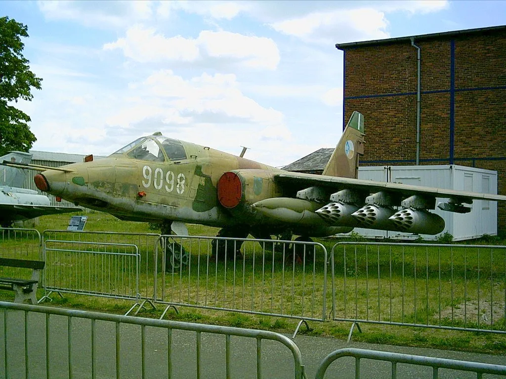 Een militair straalvliegtuig met camouflageverf en het nummer 9098, tentoongesteld buiten achter een metalen barrière.