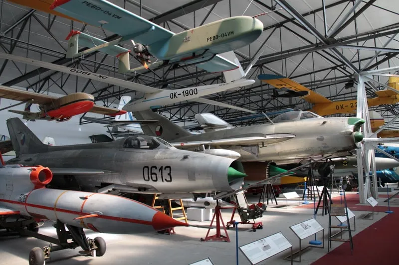 Militair vliegtuig opgesteld in een museumhangaar, waaronder een zilveren straaljager met de markering '0613' en andere vliegtuigen die aan het plafond hangen.