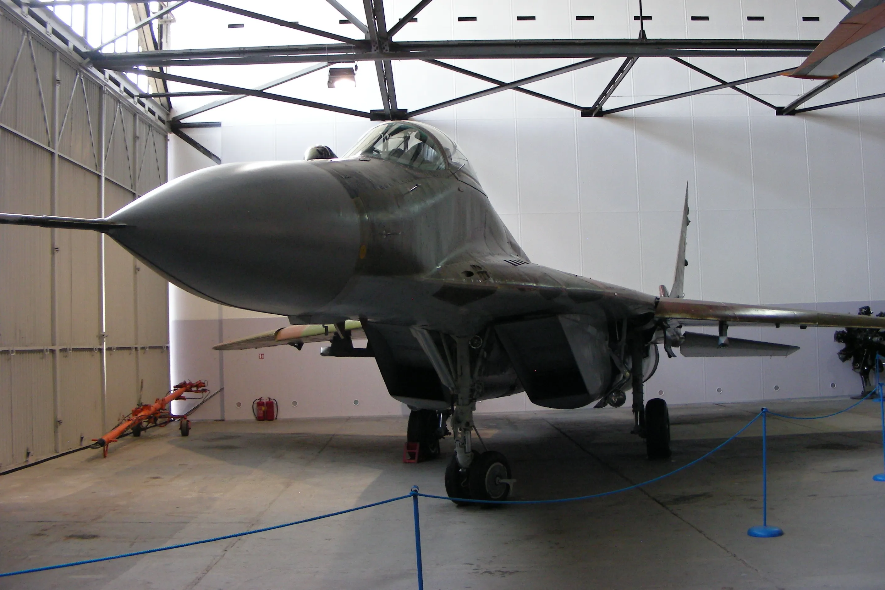 Een grijze MiG-29 straaljager tentoongesteld in een hangar met een blauw touw als afzetting ervoor