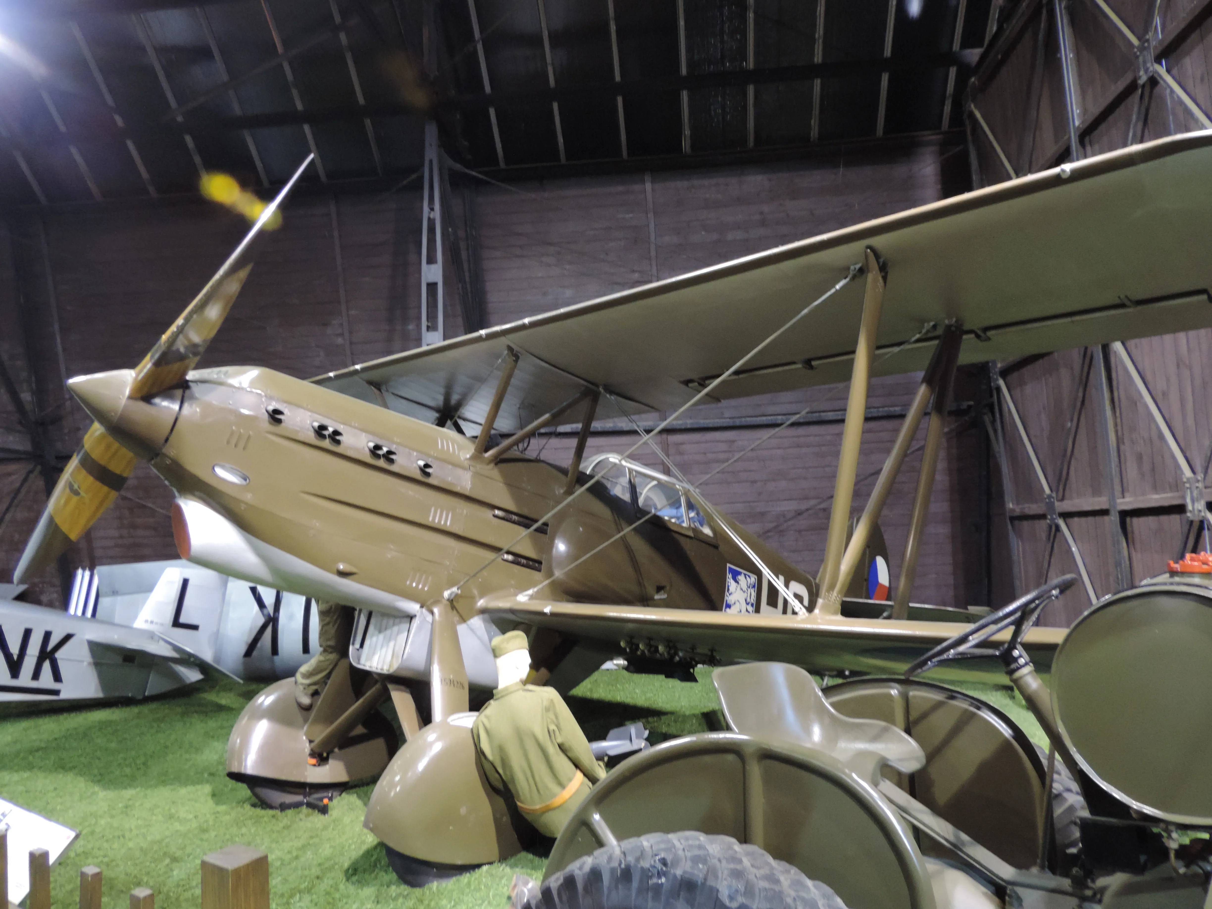 Een bruin dubbeldekker vliegtuig met een geeltippropeller en Tsjechische markeringen wordt binnenshuis tentoongesteld op kunstgras, met ander militair materieel en vliegtuigen in de buurt.