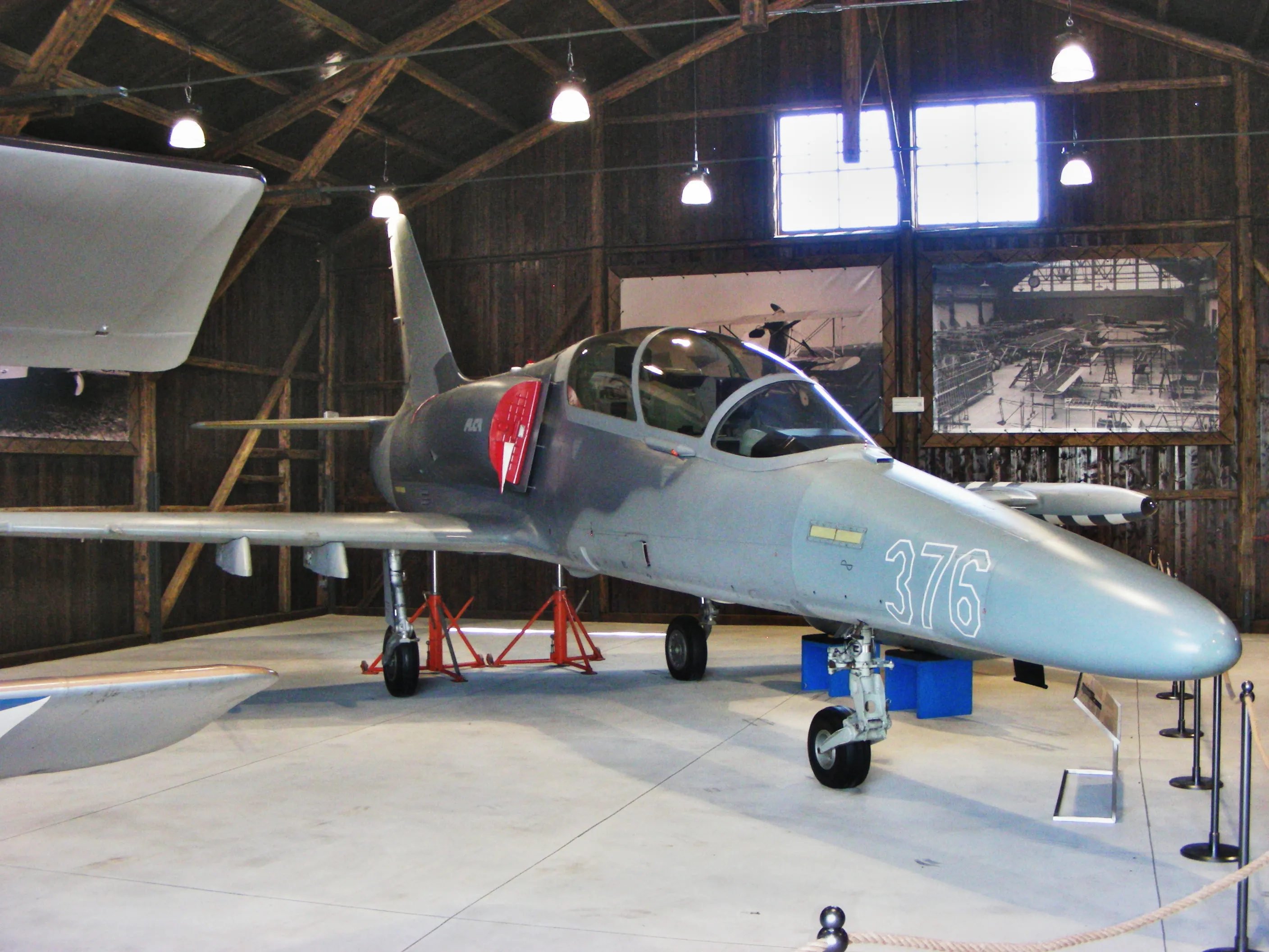 Een grijze militaire straaljager met het nummer 376 op de neus, tentoongesteld in een hangar met houten muren en industriële verlichting.