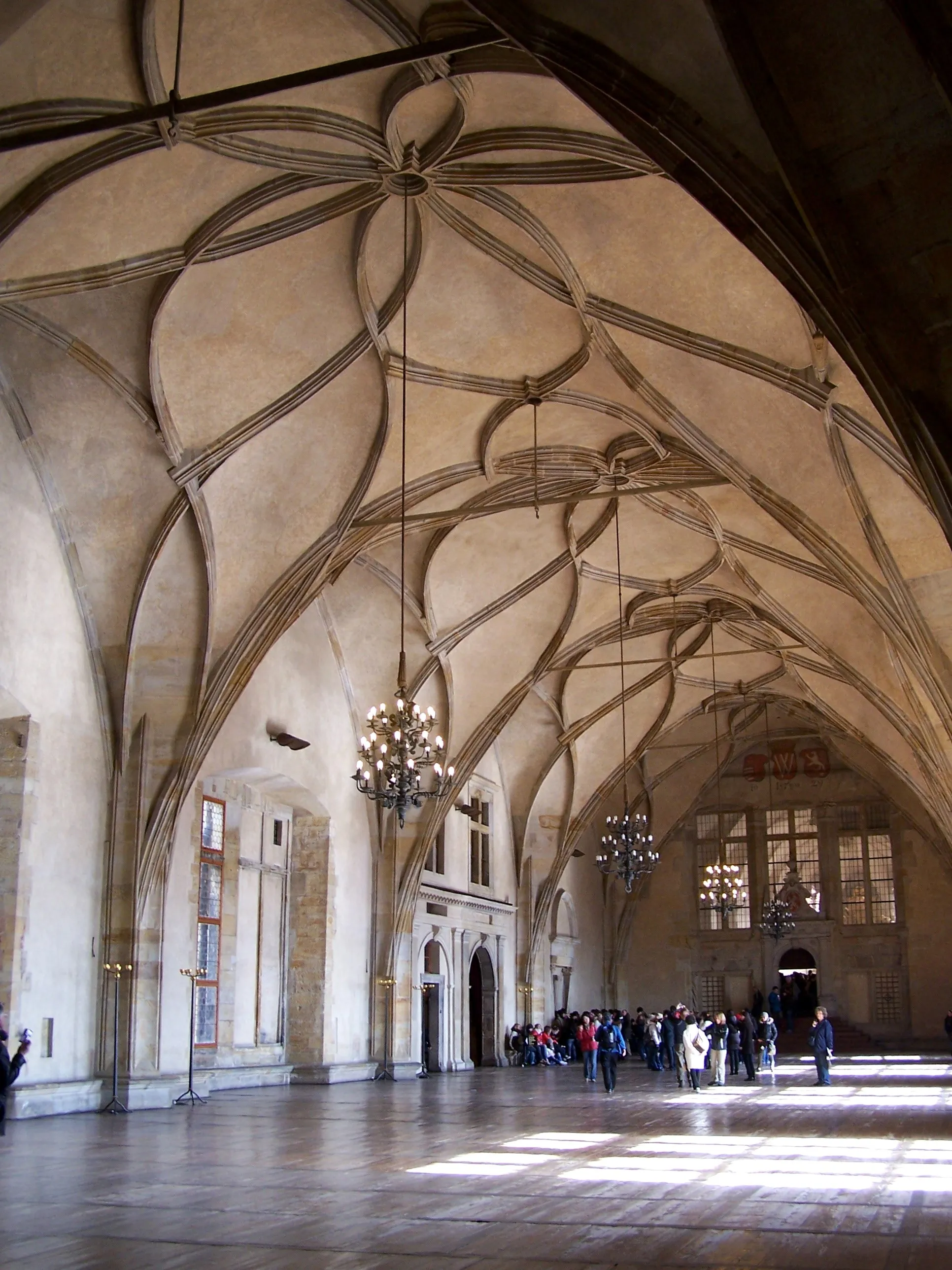 Interior of a large hall with ornate ribbed vault ceiling, chandeliers, and people walking on a polished floor.