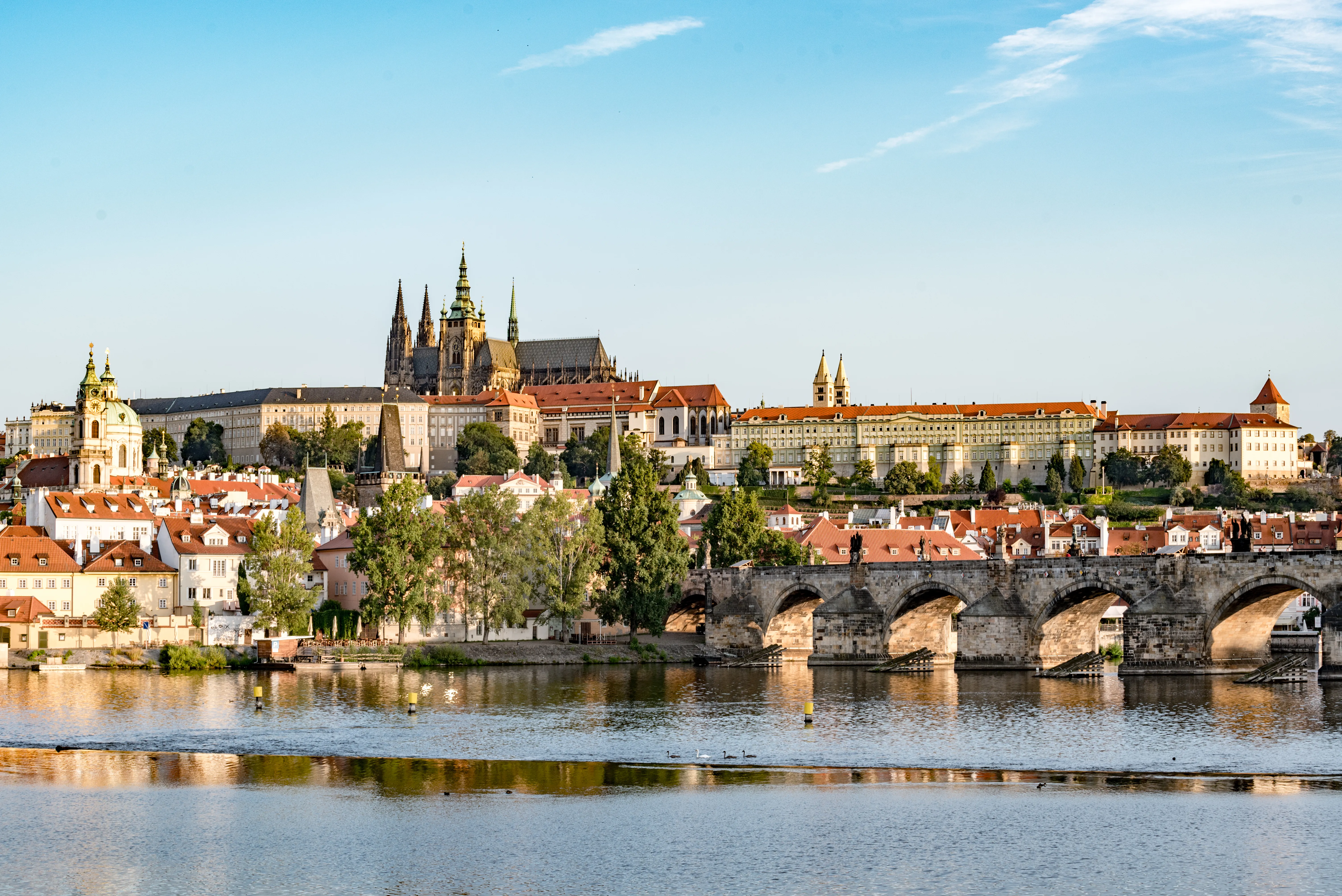 Wide panoramic view of Prague Castle complex with St. Vitus Cathedral spires rising above red-roofed buildings, seen across the Vltava River with Charles Bridge stone arches in the foreground under a blue sky.